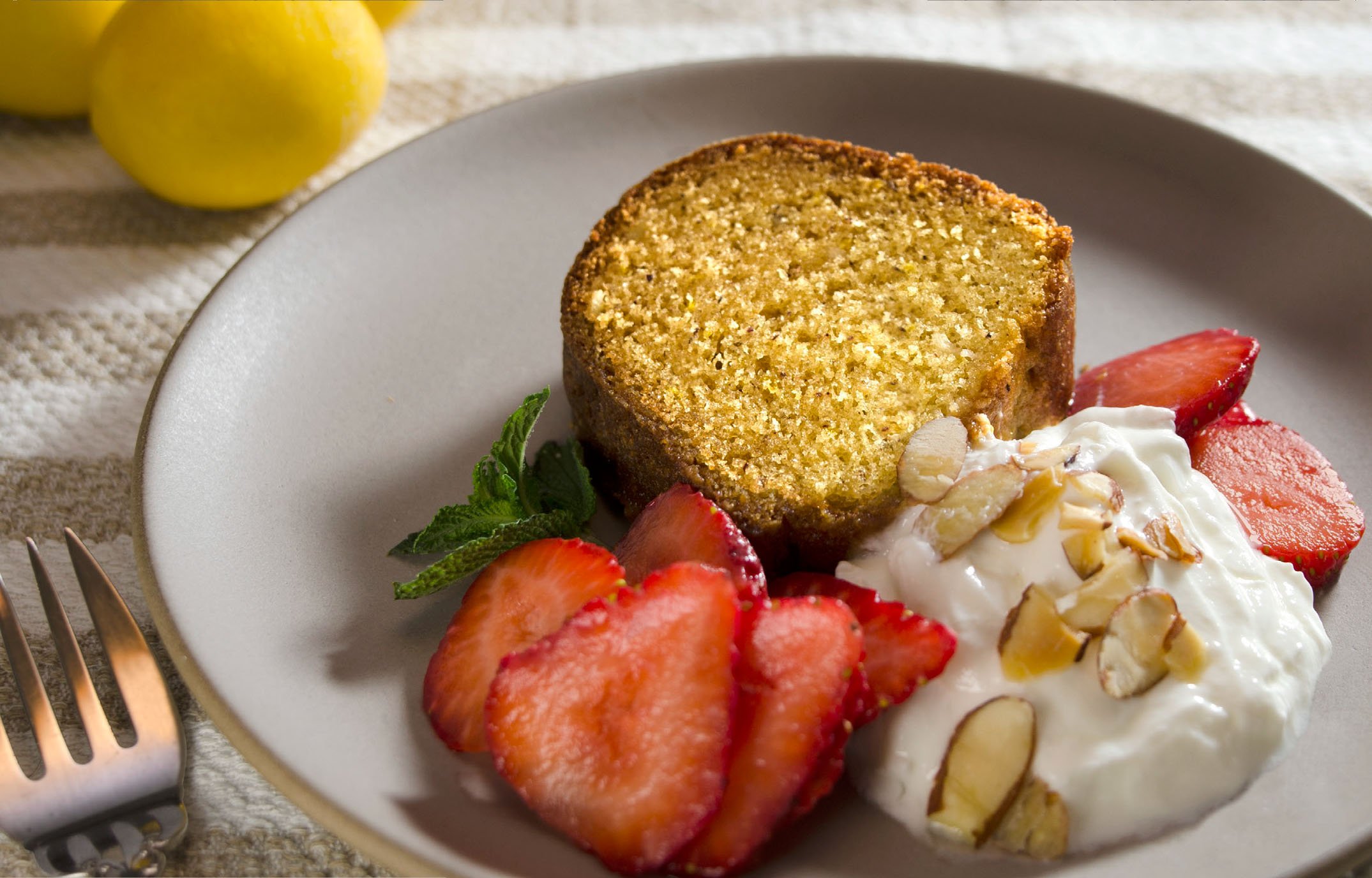 Slice of lemon pound cake served with fresh strawberries, sliced almonds, and a dollop of whipped cream garnished with a mint leaf.
