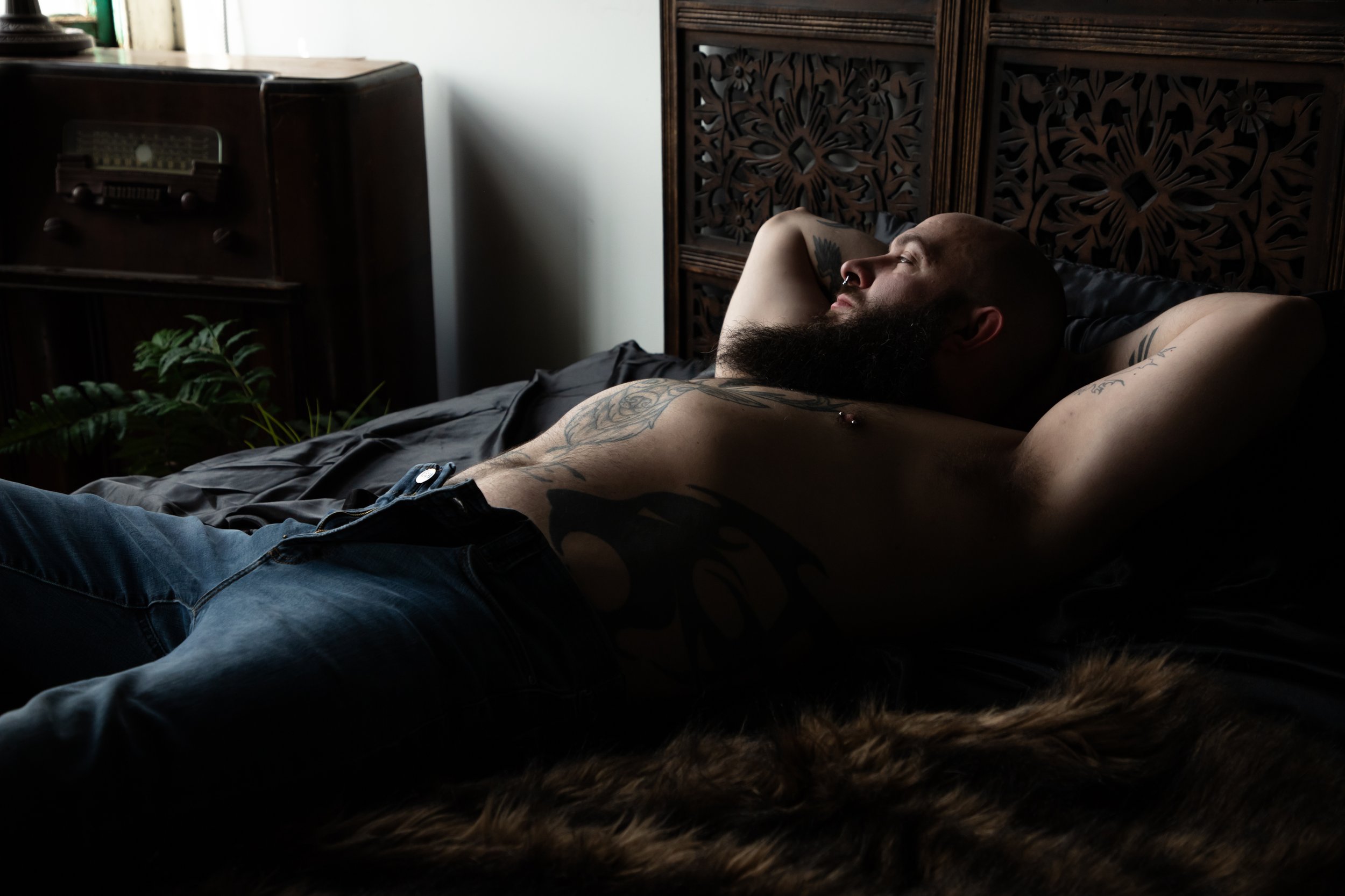 A man with a beard and tattoos relaxing on a bed with his arms behind his head in a room with wooden furniture, a plant, and a vintage radio, with natural light coming through a window.