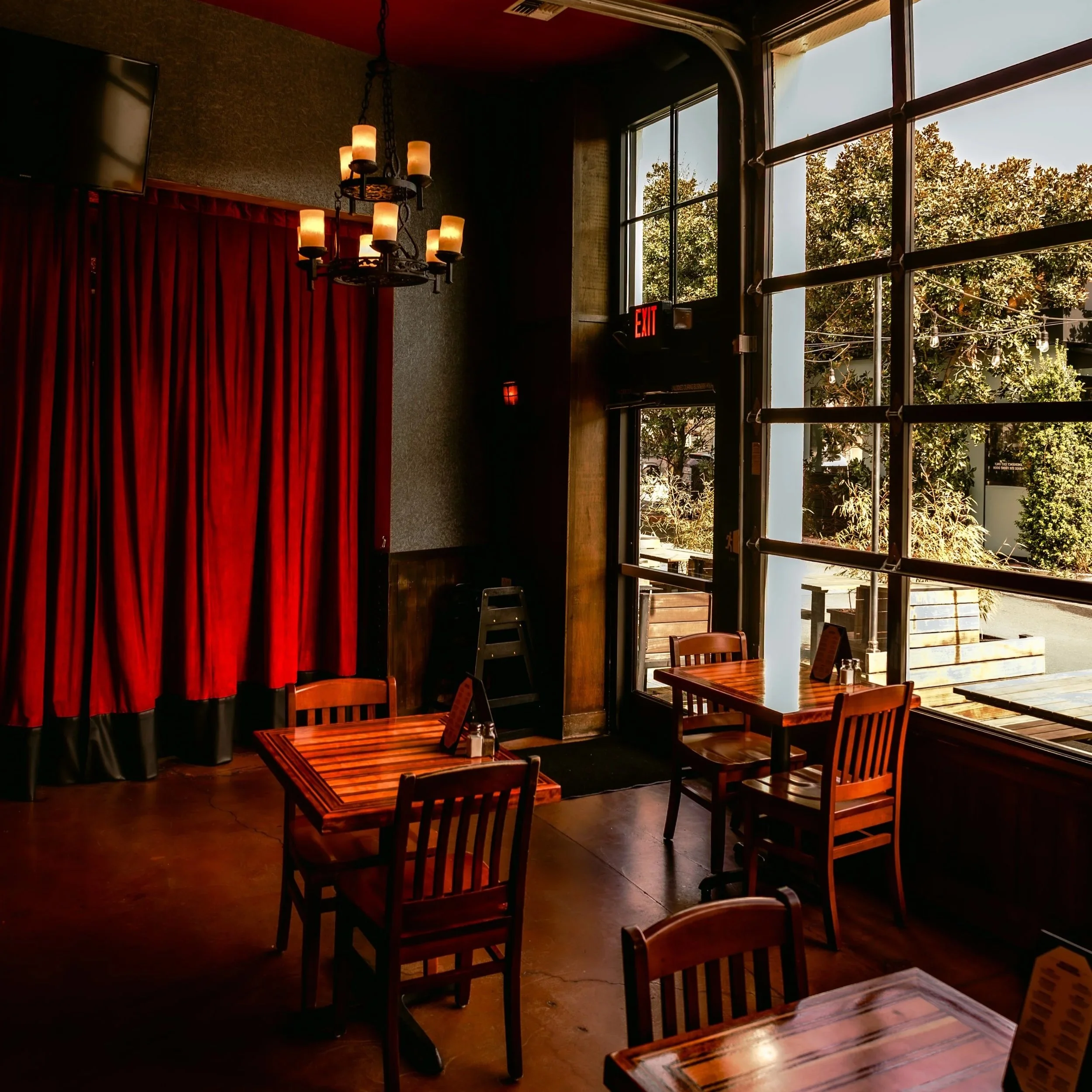 Interior of a restaurant or cafe with wooden tables and chairs, large windows letting in natural light, plants outside, a red exit sign, a chandelier with multiple lights, and a red curtain.