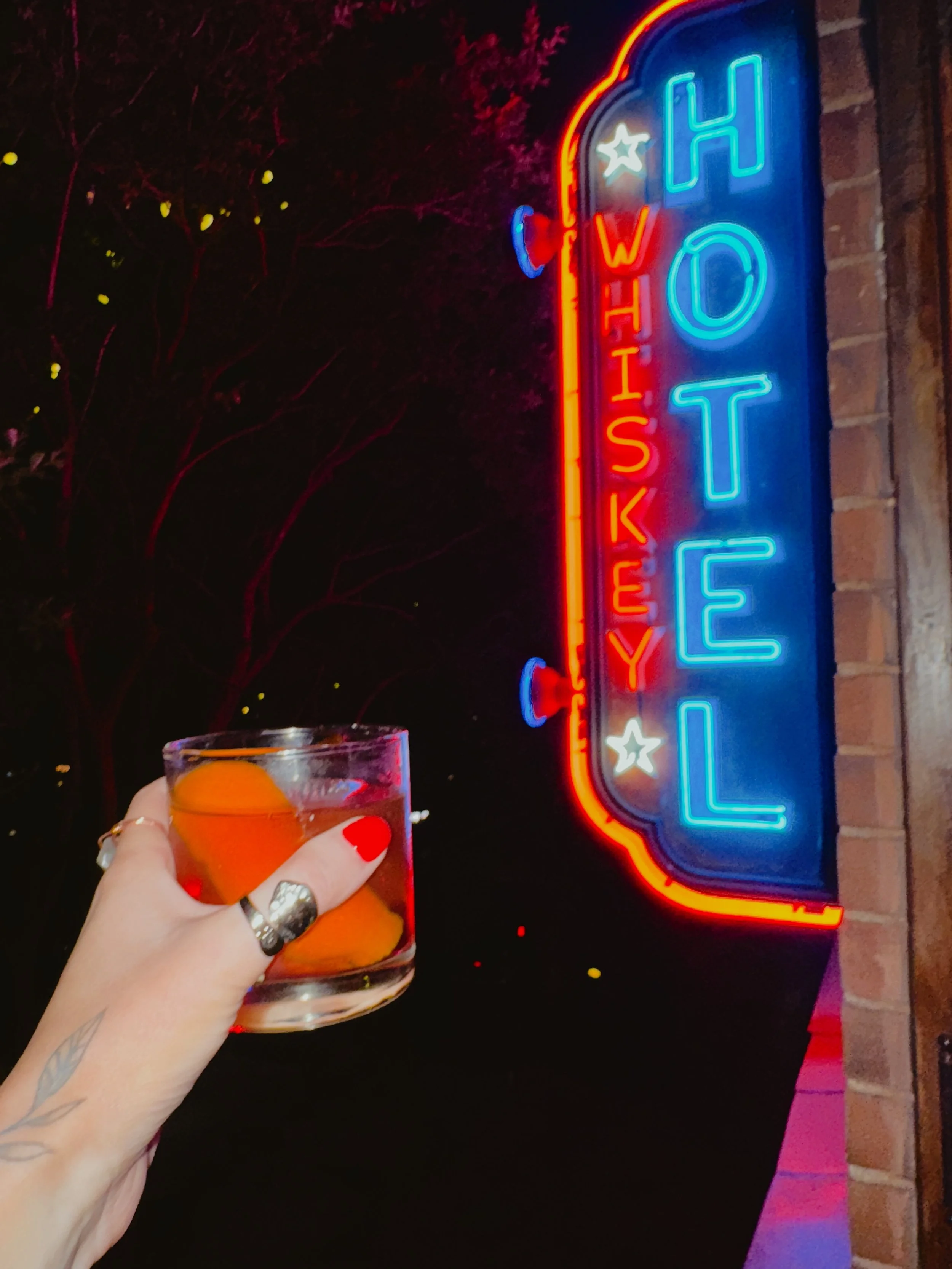 A person holding a glass with an orange slice inside, in front of a bright neon sign that spells 'HOTEL' with 'WHISKEY' vertically.