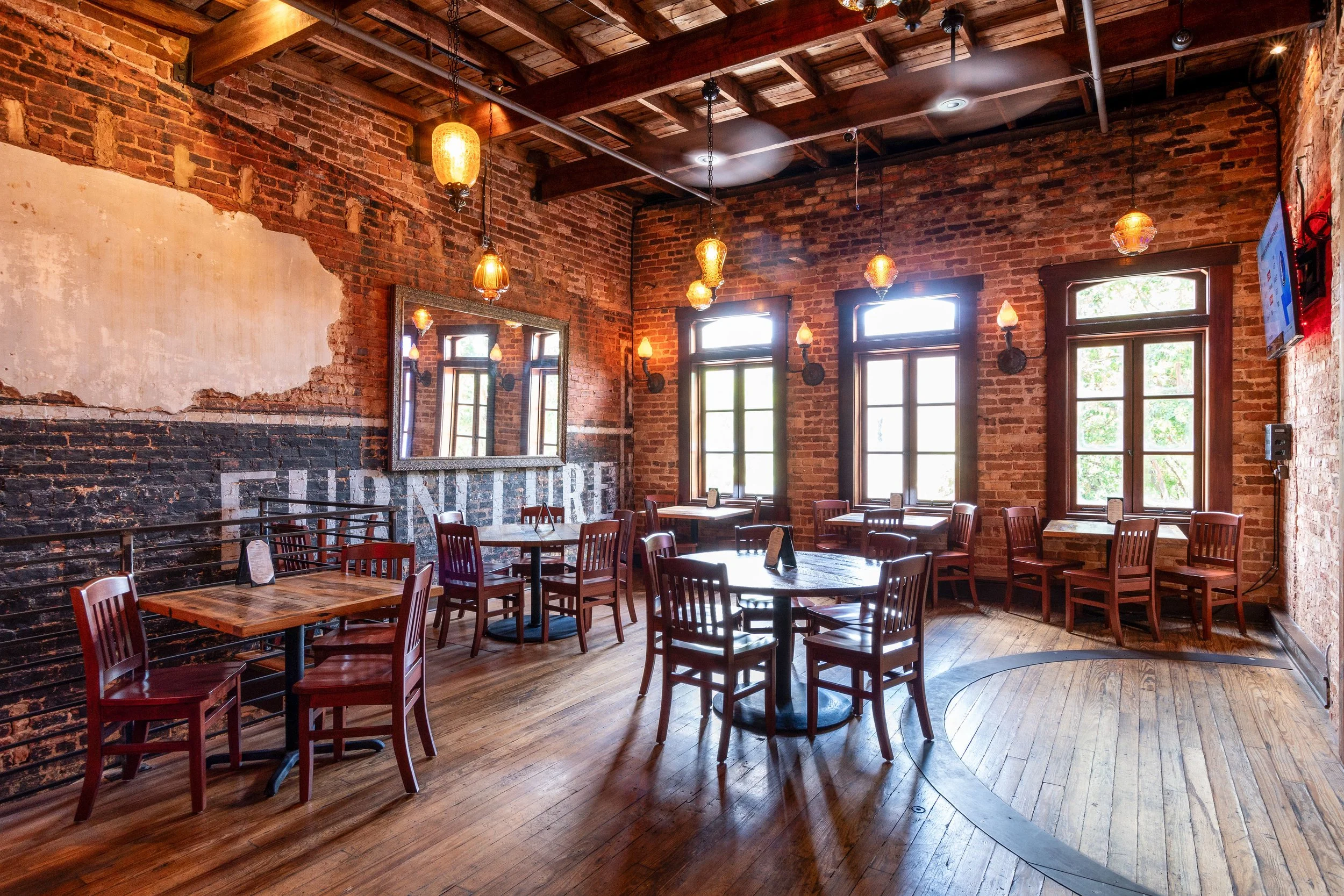 Interior of a rustic restaurant with exposed brick walls, wooden floors, multiple tables with chairs, large windows, and warm hanging pendant lights.