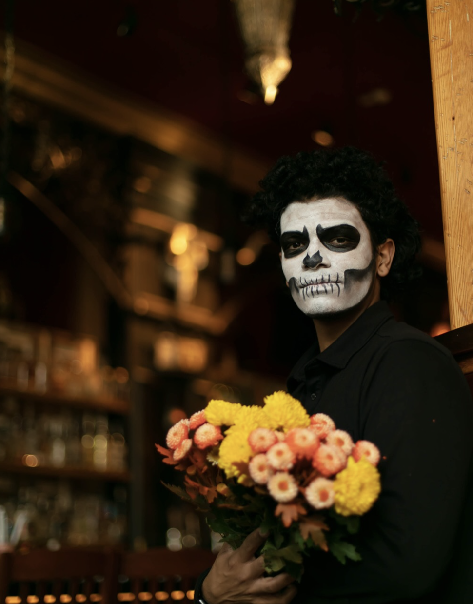 Person with black curly hair and skeleton face paint holding a bouquet of yellow and pink flowers indoors.