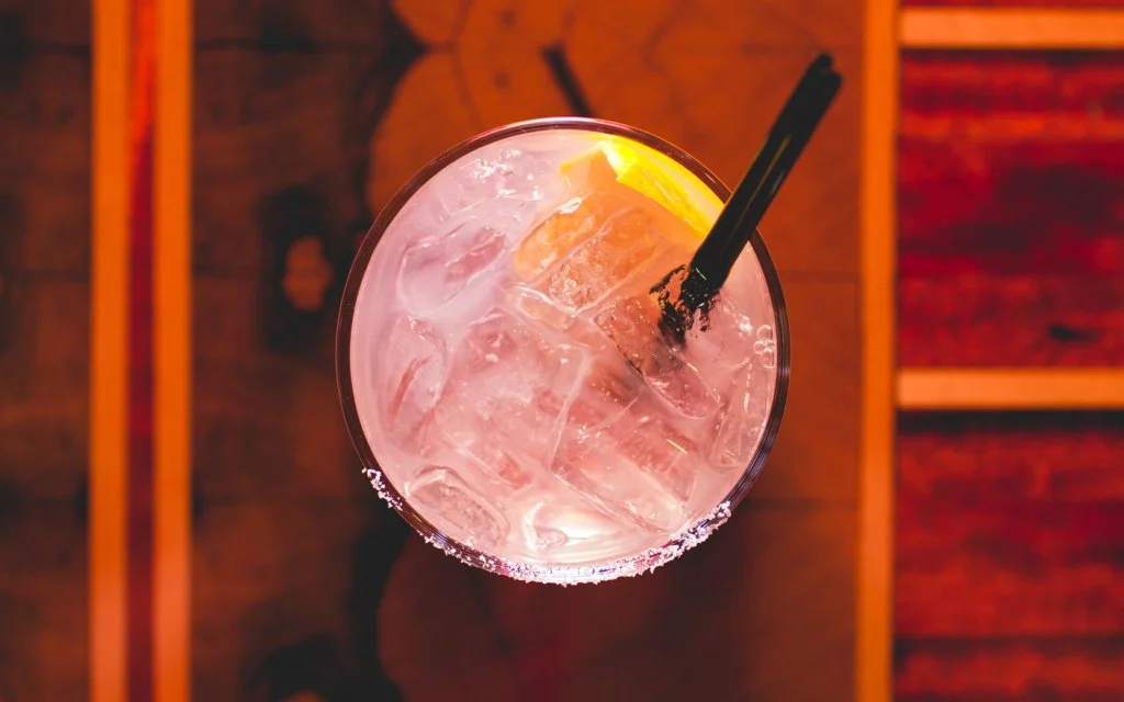 Top view of a pink-colored cocktail with ice cubes and a lemon wedge, with two black straws.