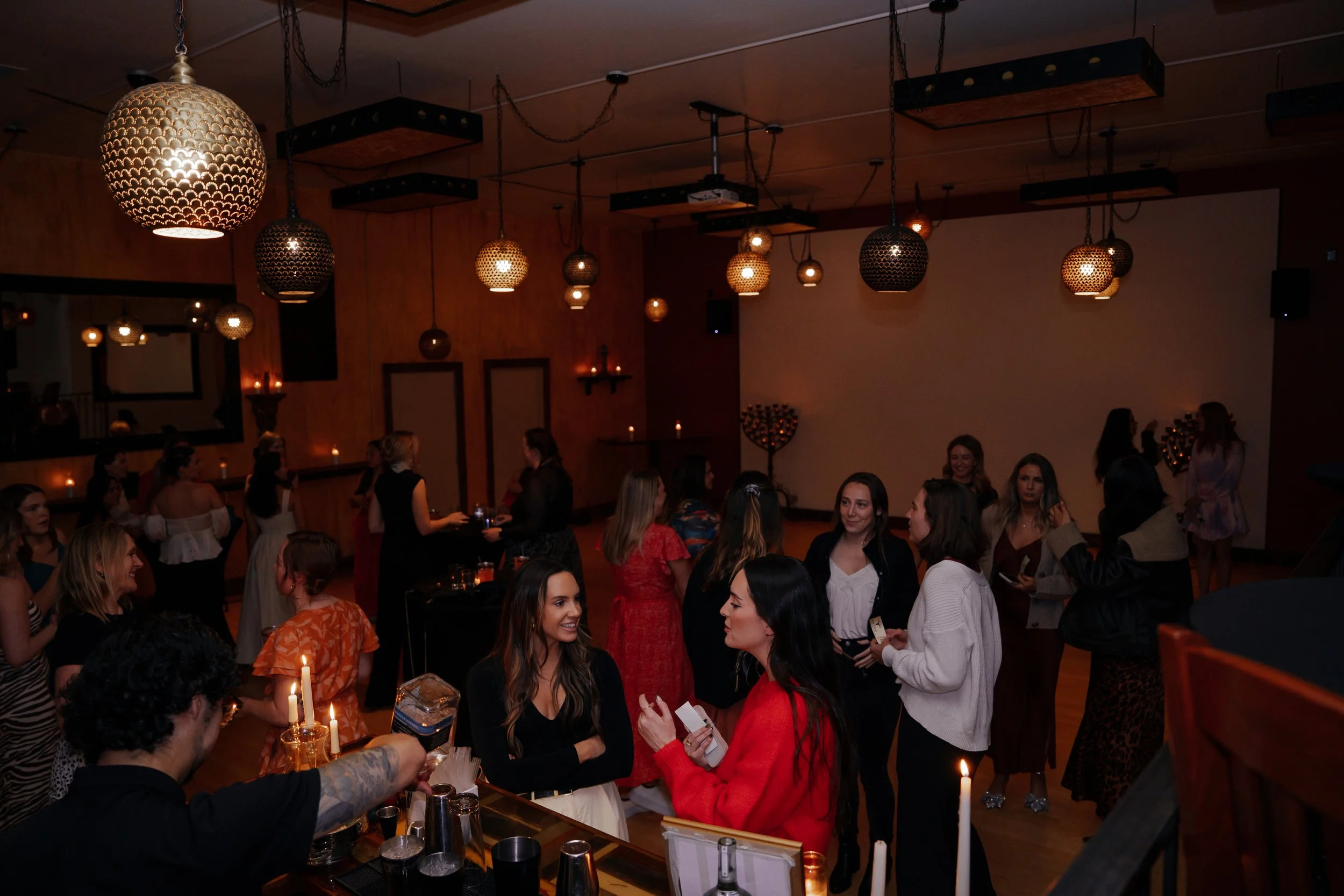Group of women socializing at a party in a warmly lit venue with hanging pendant lights and candles.
