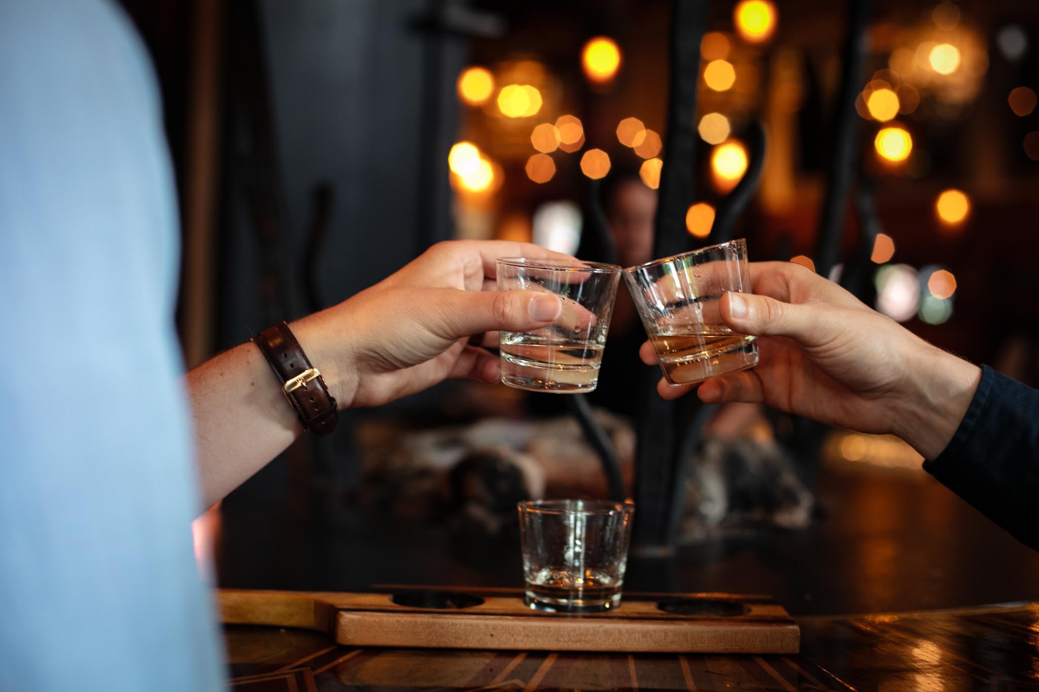 Two people clinking glasses of whiskey in a dimly lit bar with warm, blurred lights in the background.