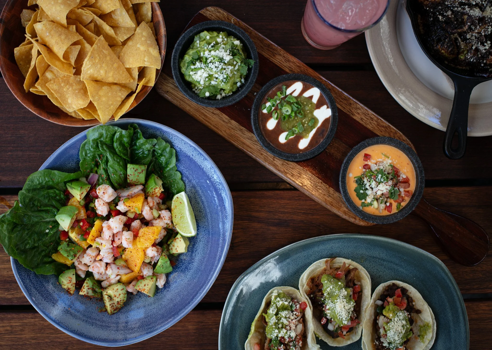 A variety of Mexican dishes on a wooden table, including a bowl of tortilla chips, a seafood salad with avocado and lemon, three small bowls of different salsas or sauces, and three tacos with meat, vegetables, and cheese.