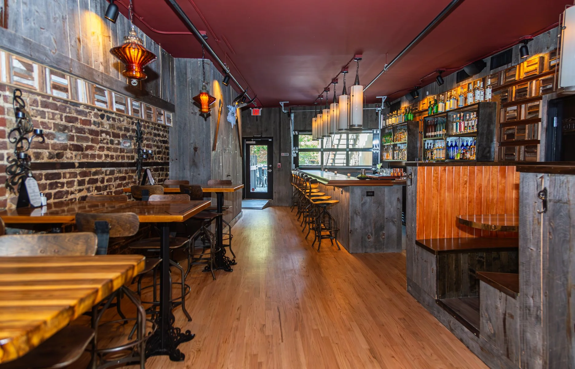 Interior of a rustic-style bar with wooden floors, brick and wood walls, and a bar counter with barstools. Decor includes hanging pendant lights and rustic ornaments.