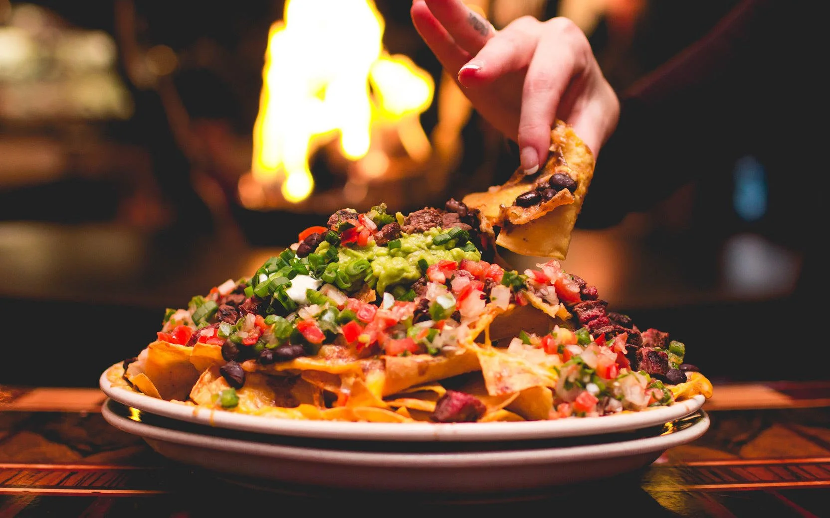 A hand holding a tortilla chip dipped into loaded nachos topped with cheese, black beans, guacamole, salsa, sour cream, and chopped green onions, with a blurry background and warm fire.