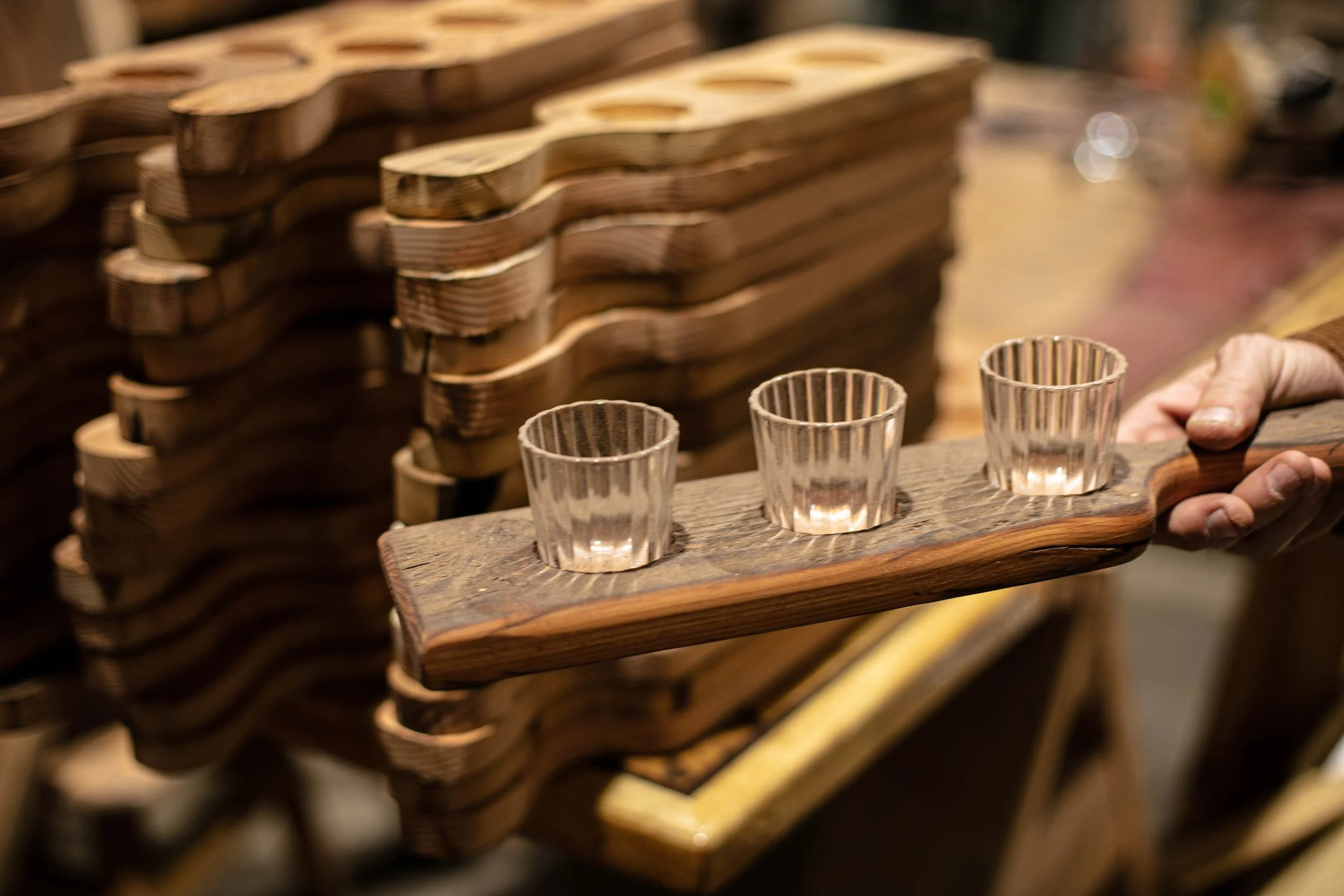 A person holding a wooden tray with three glass shot glasses, with stacks of wooden trays in the background.