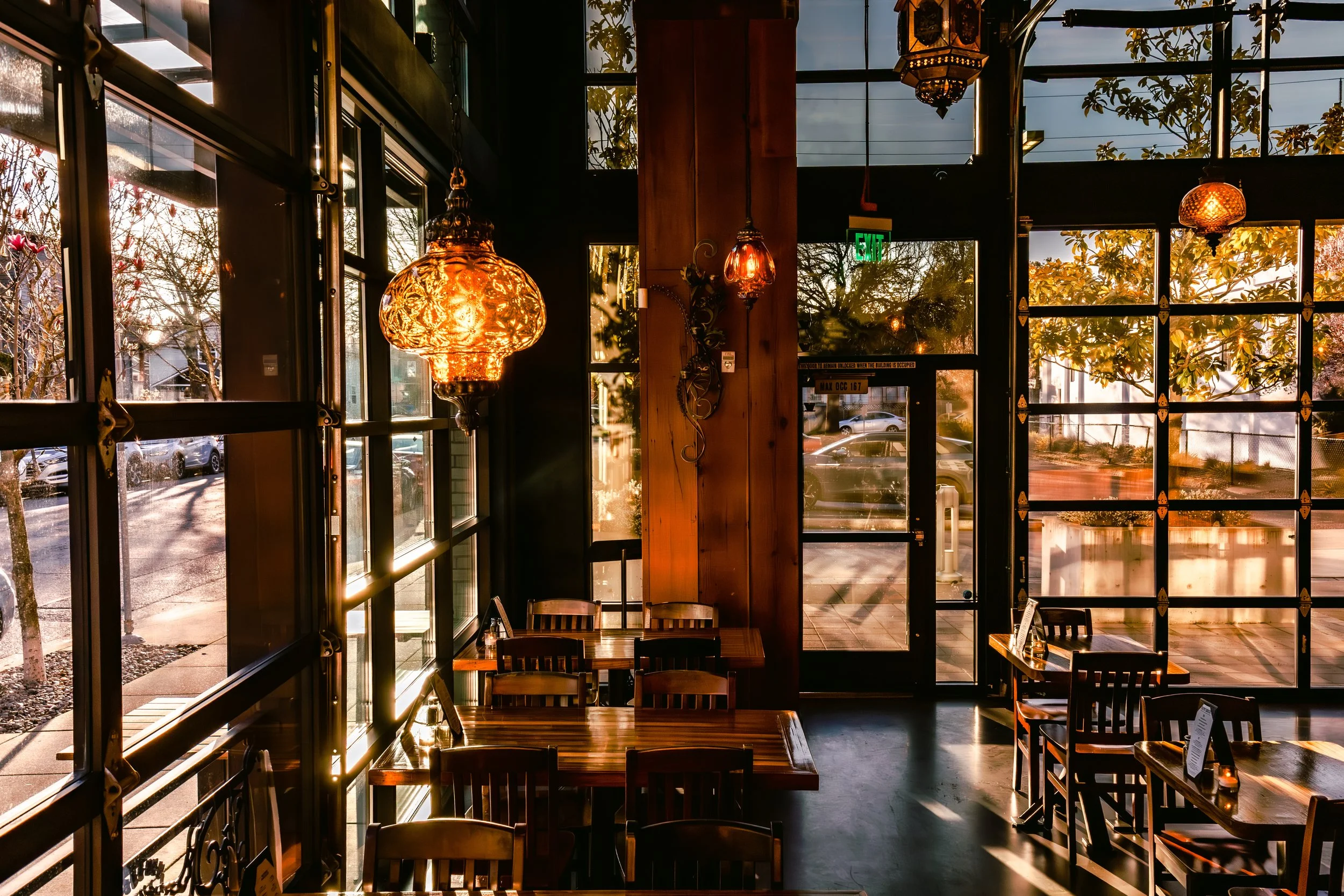 Interior of a cozy café with wooden tables and chairs, illuminated by warm decorative hanging lights. Large windows reveal trees and parked cars outside, suggestive of late afternoon lighting.