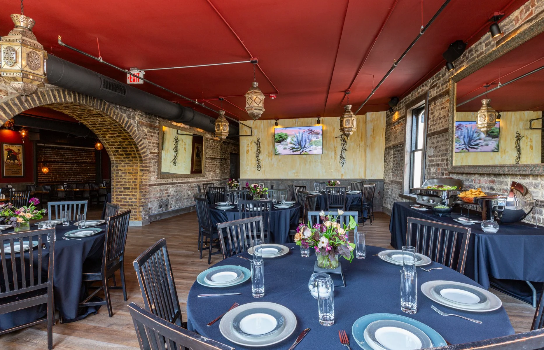 Interior of a restaurant with brick walls, wooden floors, and round tables covered with dark blue tablecloths, set with plates, glasses, and silverware, decorated with floral centerpieces, and large windows allowing natural light.
