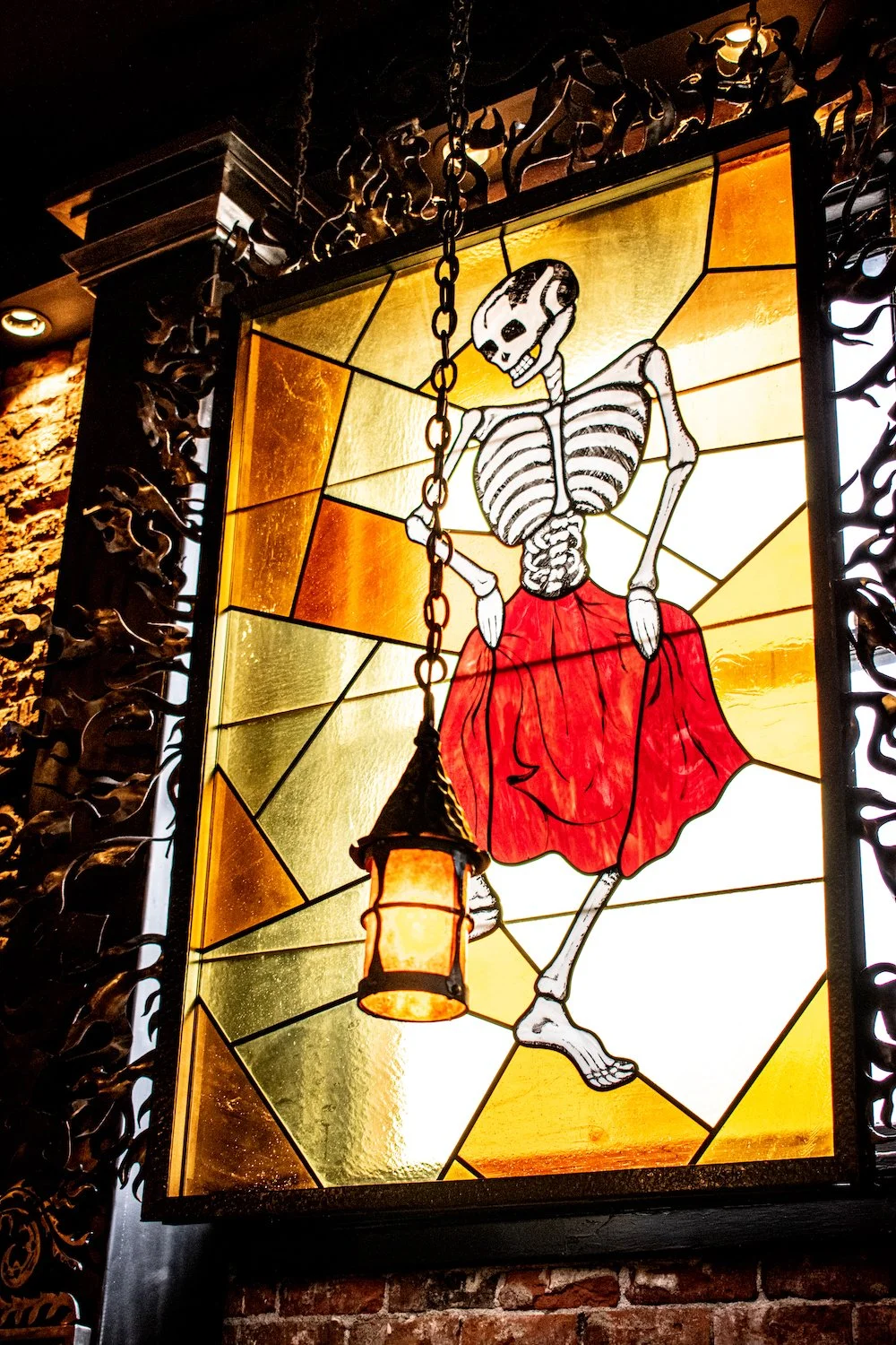 A stained glass window depicting a skeleton wearing a red skirt with a lantern hanging from its hand.