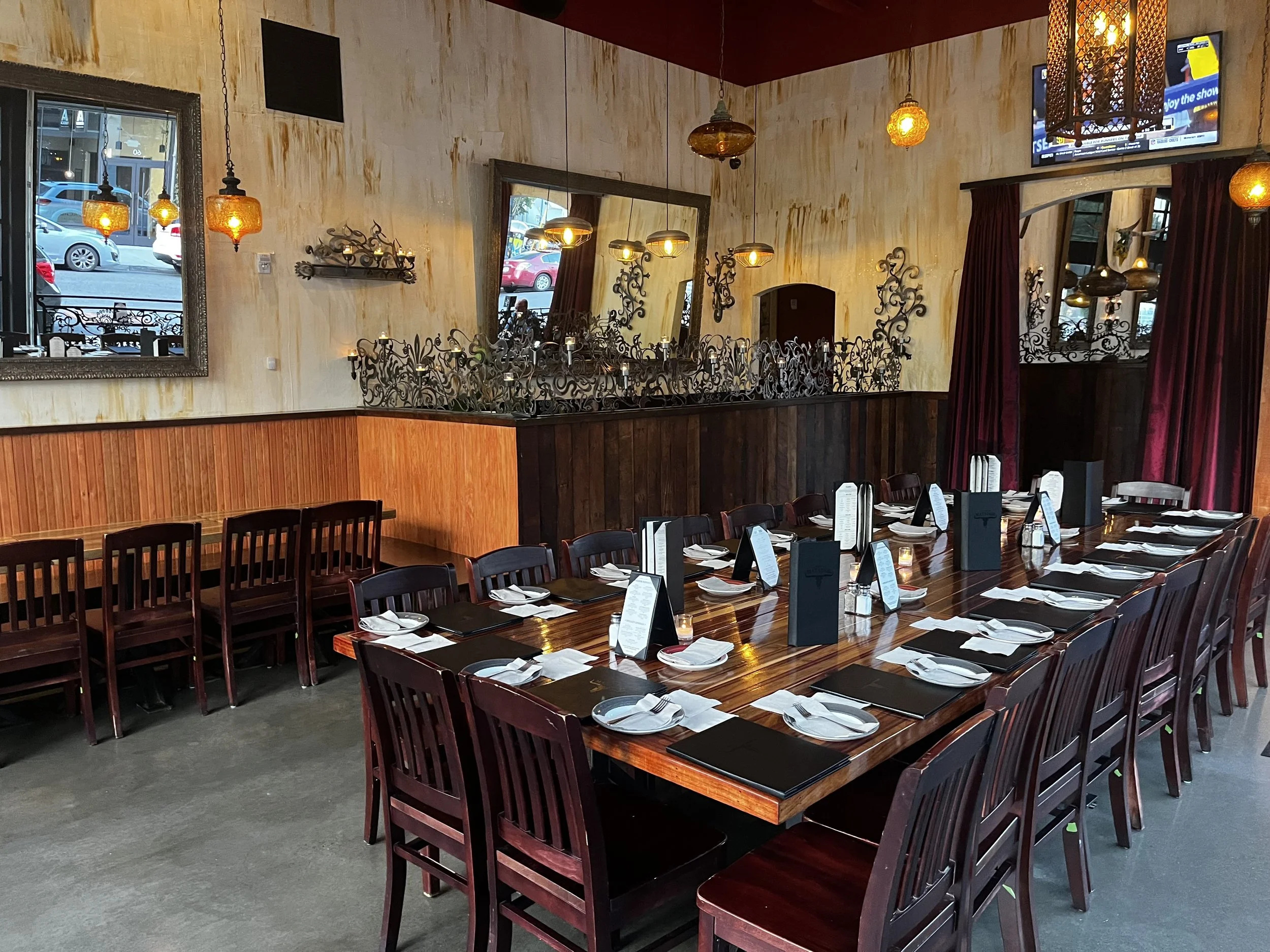 A restaurant dining area with a long wooden table set for a large group, each place with white napkins, silverware, a menu, and a glass. The room has warm lighting, dark wood accents, a textured beige wall, mirrors, ornate black ironwork, and hanging pendant lights with yellowish glow. There are large windows with red curtains.