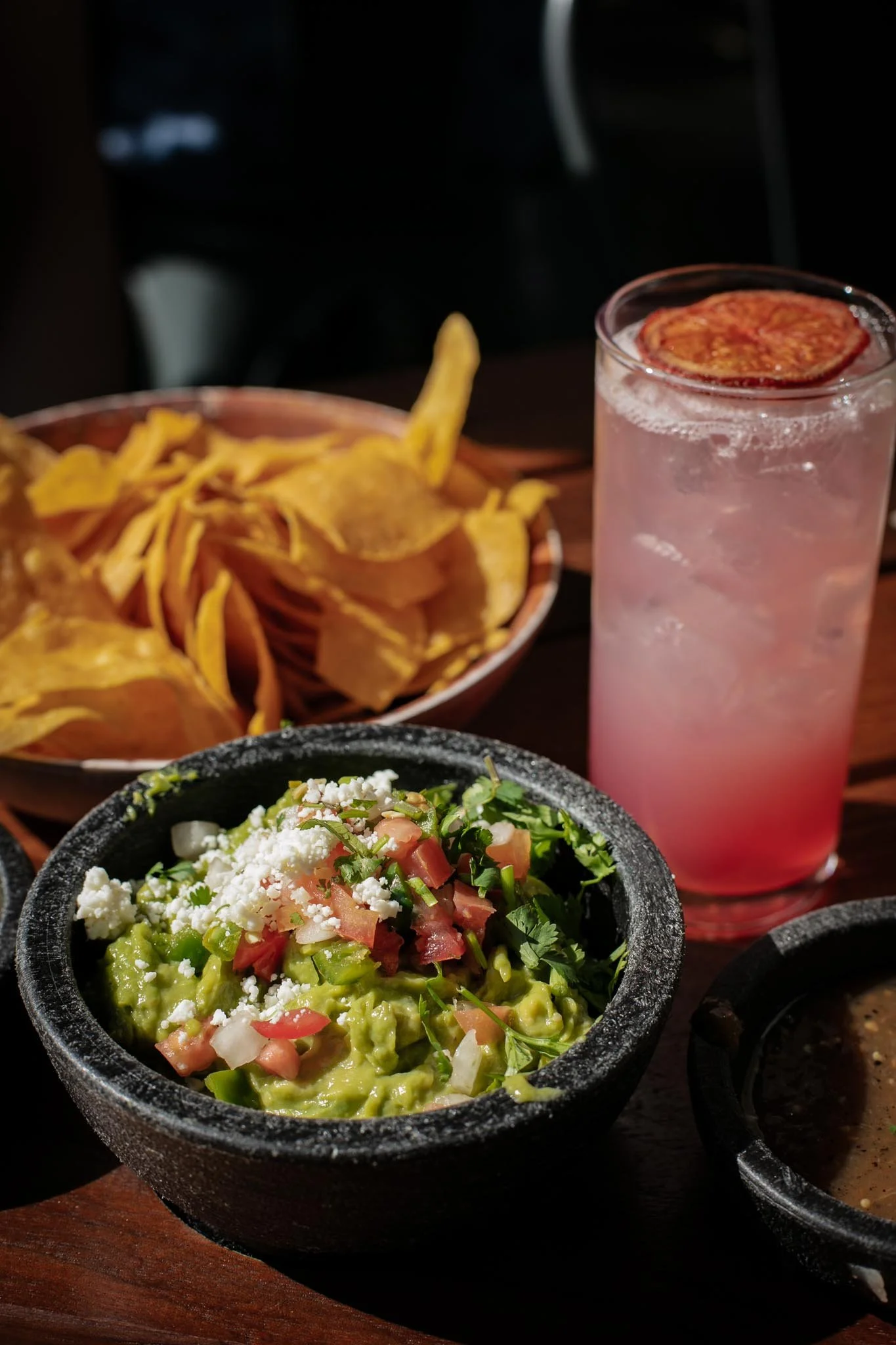 A bowl of guacamole with chopped tomatoes, onions, cilantro, and crumbled cheese, alongside a plate of yellow tortilla chips, and a pink drink with a slice of dried lime.