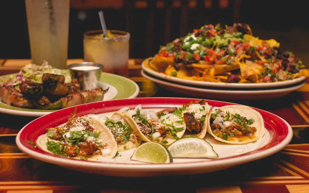 Plate of three tacos topped with chopped onions, cilantro, and cheese, garnished with lime wedges. In the background, nachos topped with meat, cheese, lettuce, tomatoes, and other toppings. Two glasses of drinks are also visible.
