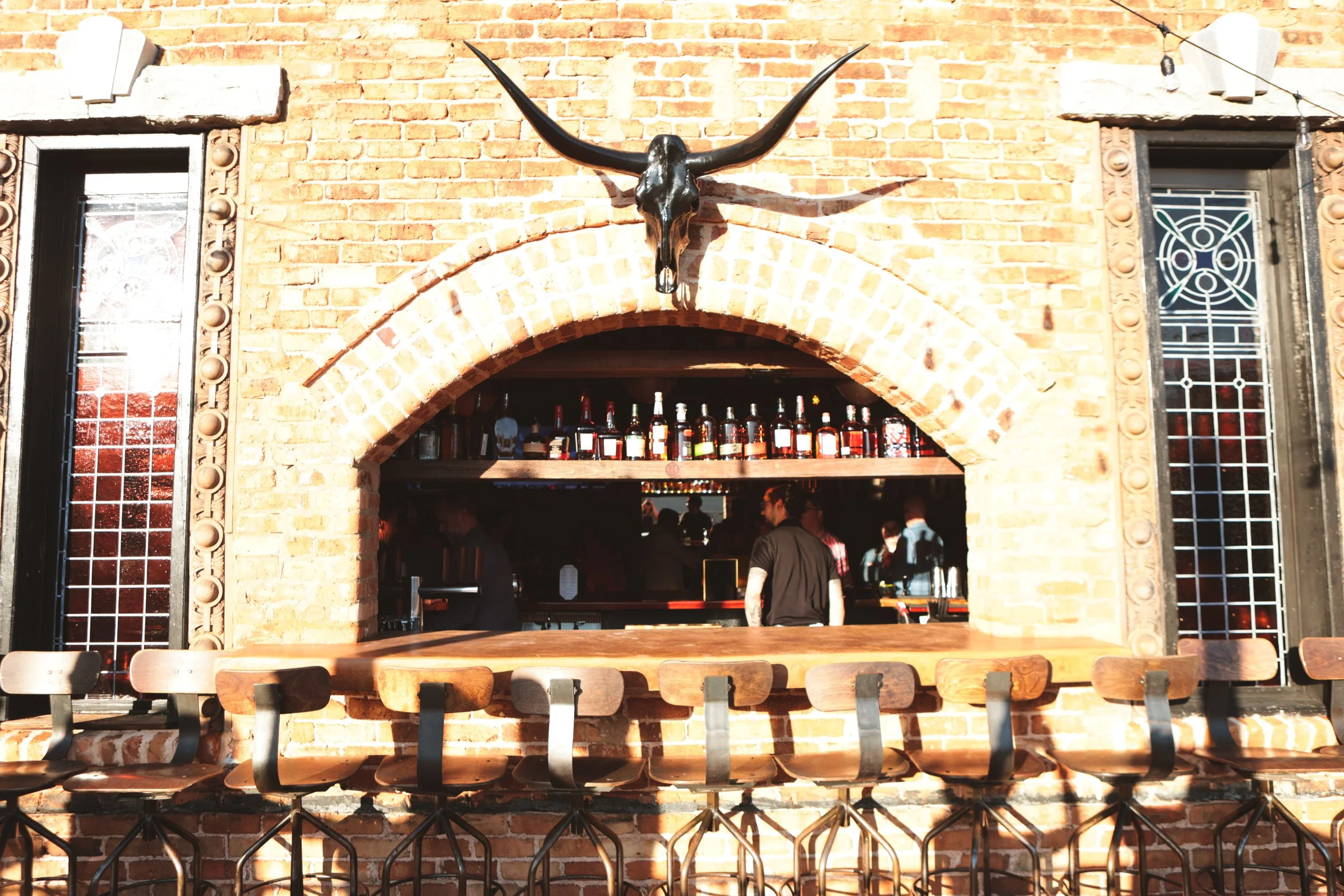 A brick wall with a mounted animal skull and horns with a bar inside a restaurant or bar. Several bottles of alcohol are lined up on a shelf behind the bar, and people are seen inside the establishment.