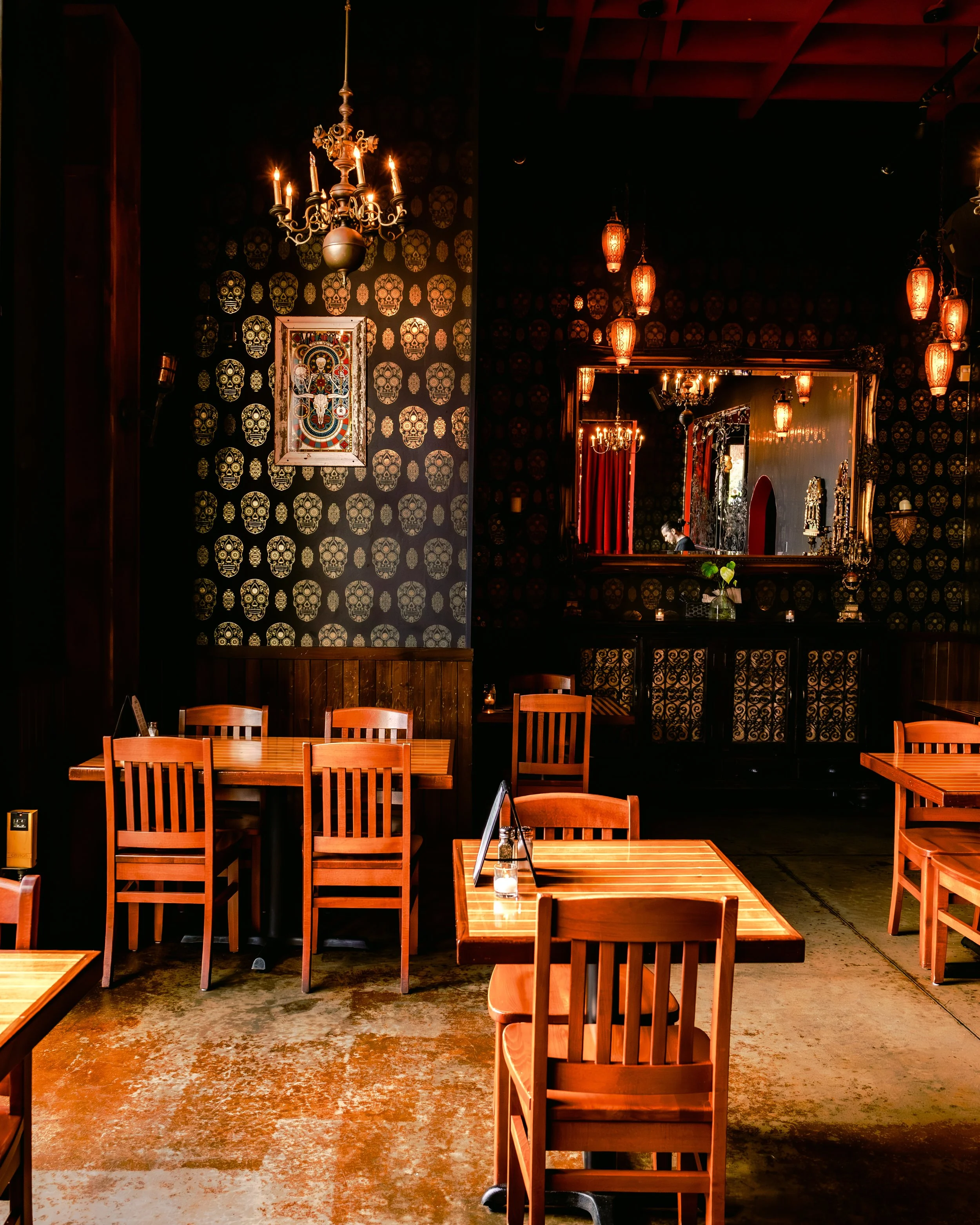 A cozy restaurant interior with wooden tables and chairs, dark patterned wall with gold skull designs, ornate chandelier, and hanging lanterns reflecting in a large mirror. A person is visible in the mirror, possibly a staff member.