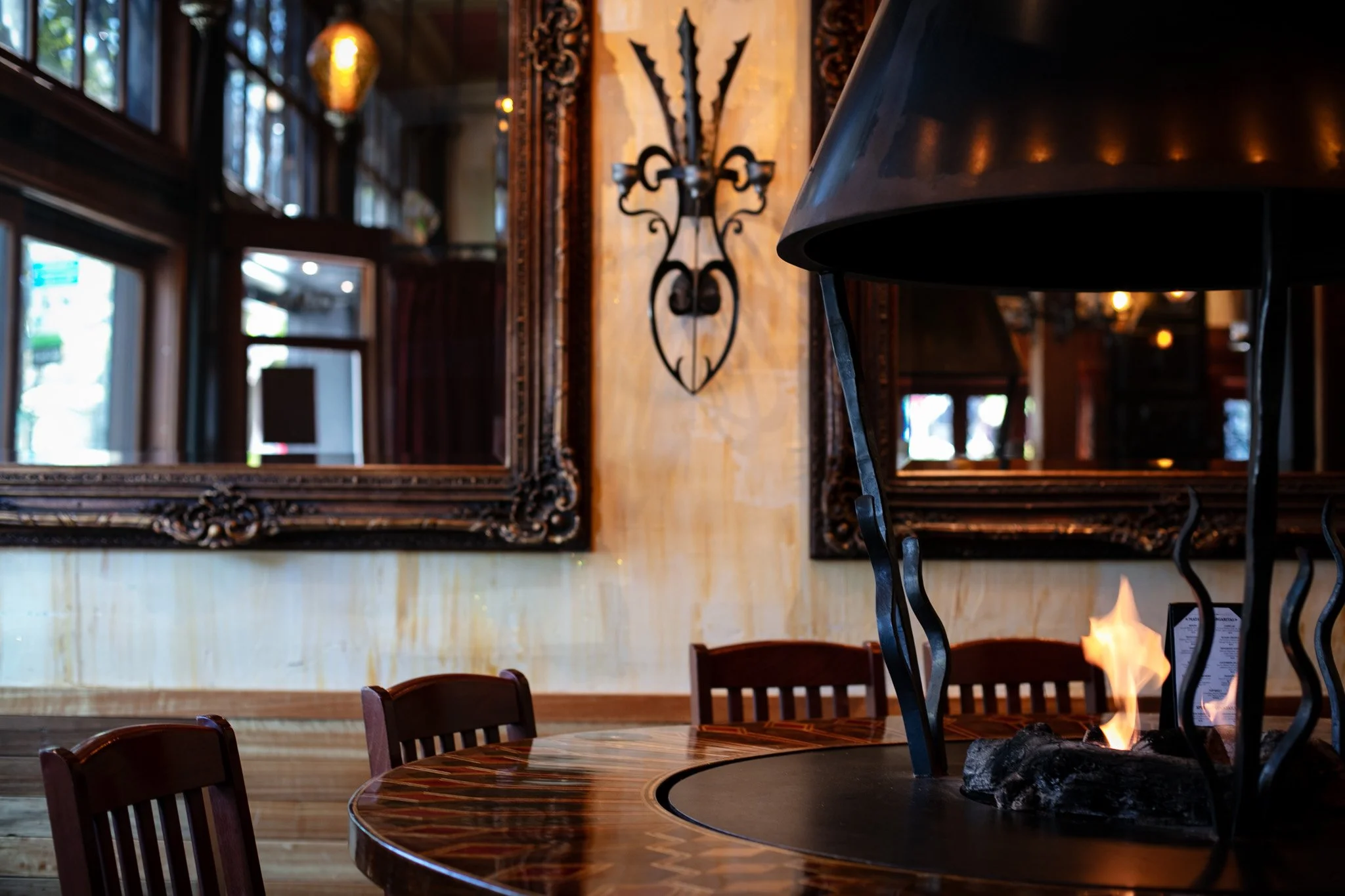 Interior of a restaurant or cafe with a round wooden table, a fireplace, and ornate framed mirrors on the wall. Reflections of windows and lights are visible in the mirrors.