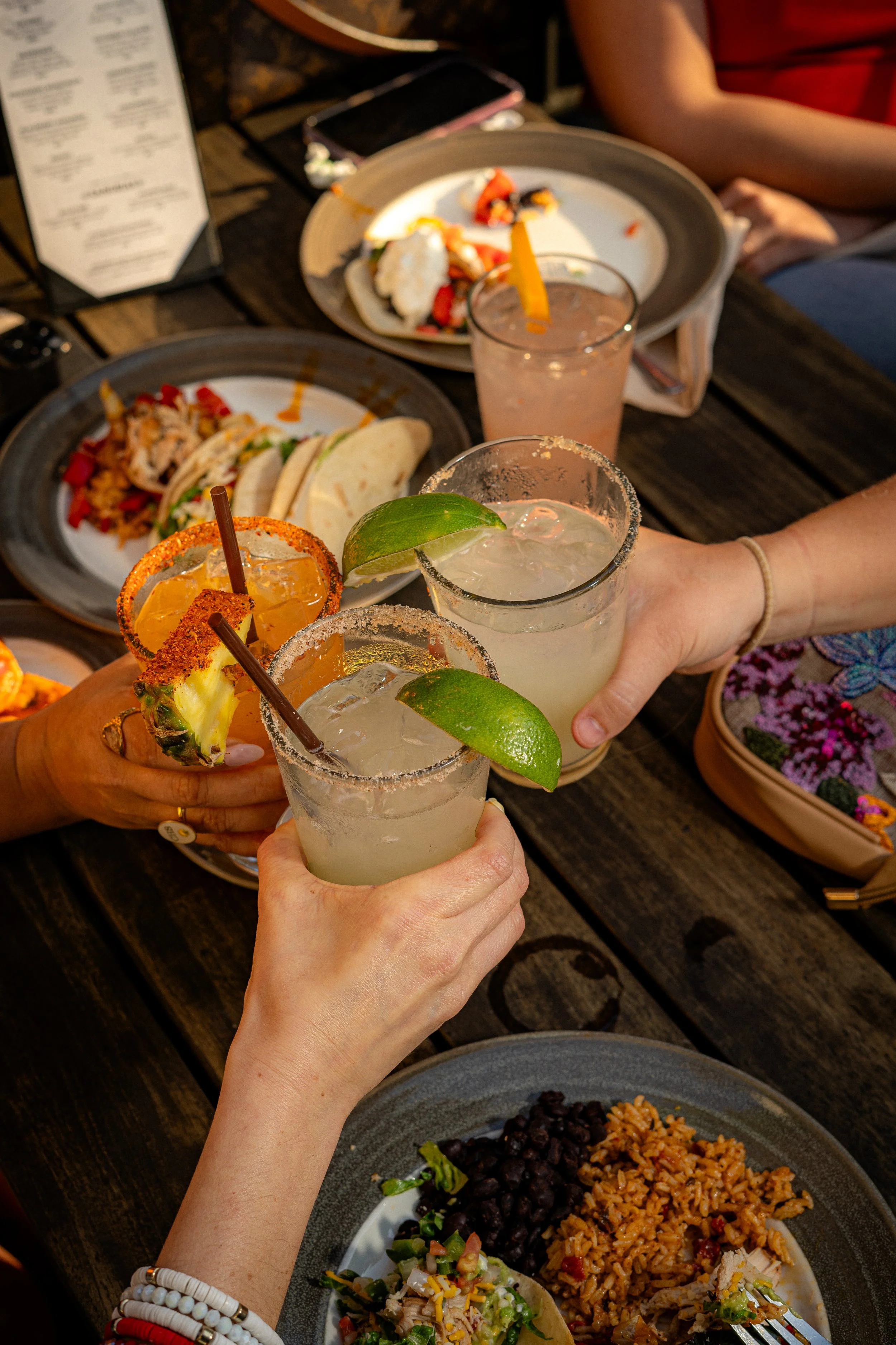 People clinking glasses of margaritas at a table with food, including tacos, rice, beans, and plates with remnants of food, in a casual dining setting.