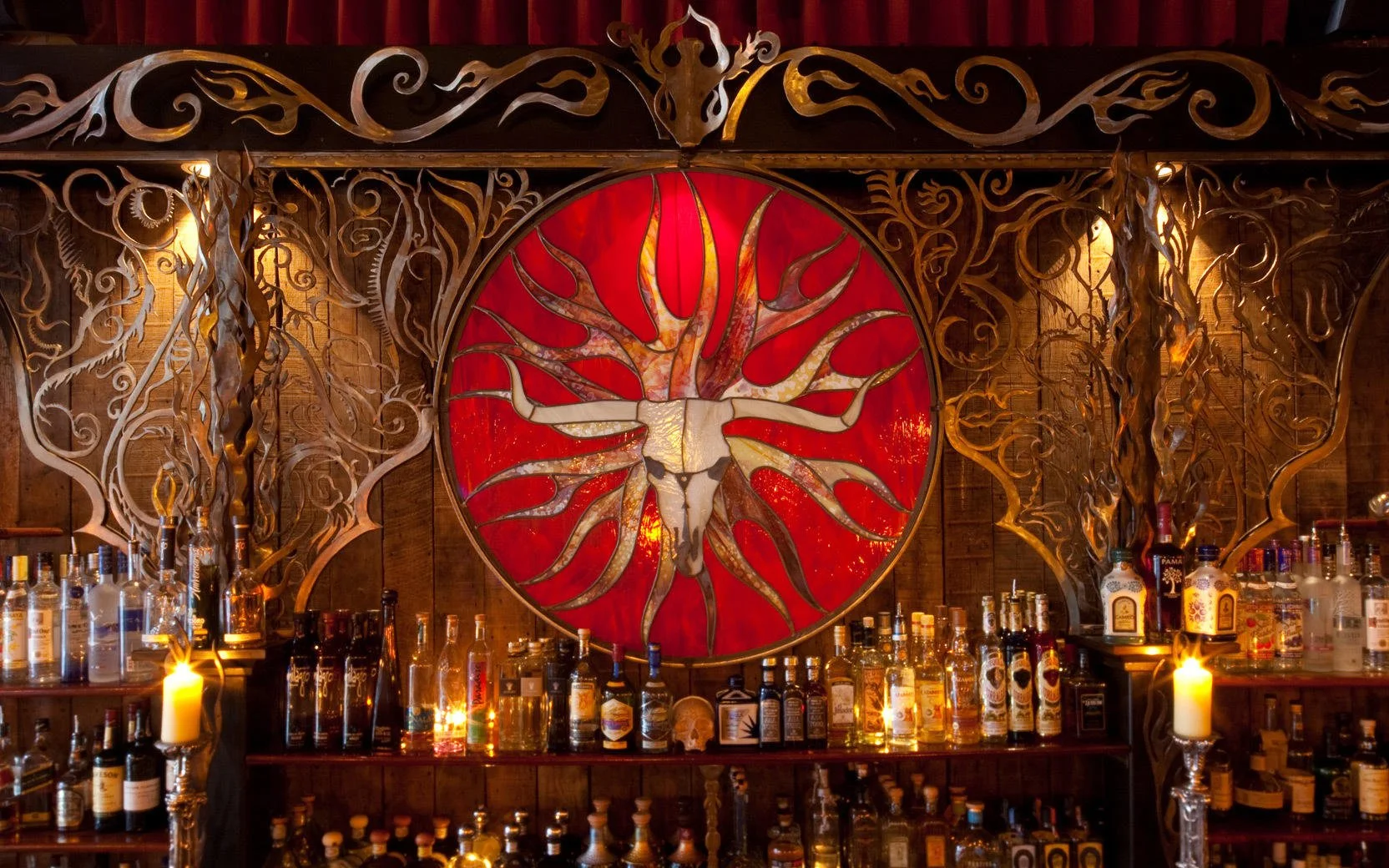 Decorative bar display with a large circular metal art piece of a bull skull with pink and orange antlers on a red background, surrounded by ornate metalwork and bottles of alcohol.
