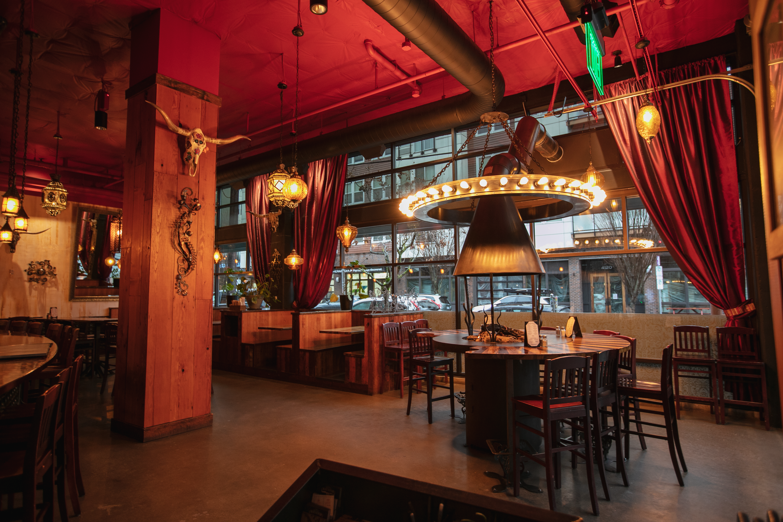 Interior of a restaurant with wooden beams, red curtains, hanging lanterns, and a large round fixture with lights. Large windows show parked cars outside.
