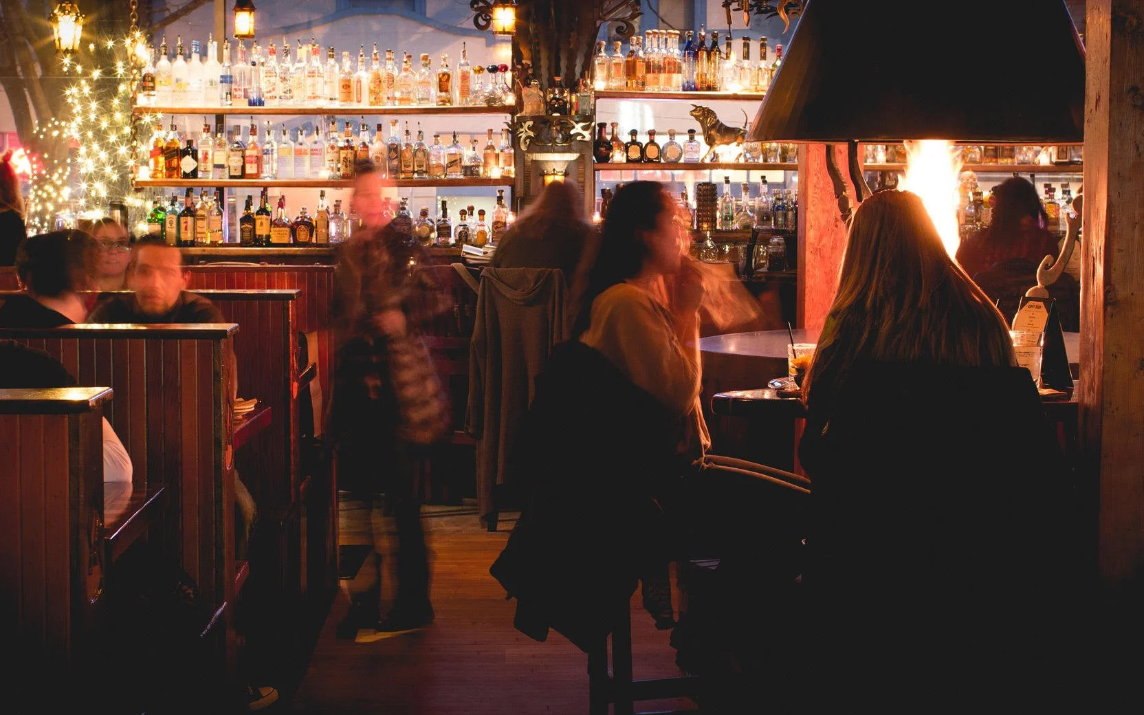 People sitting and standing in a cozy bar or restaurant with a well-stocked liquor shelf in the background. A fire is burning in a fireplace, creating a warm atmosphere.