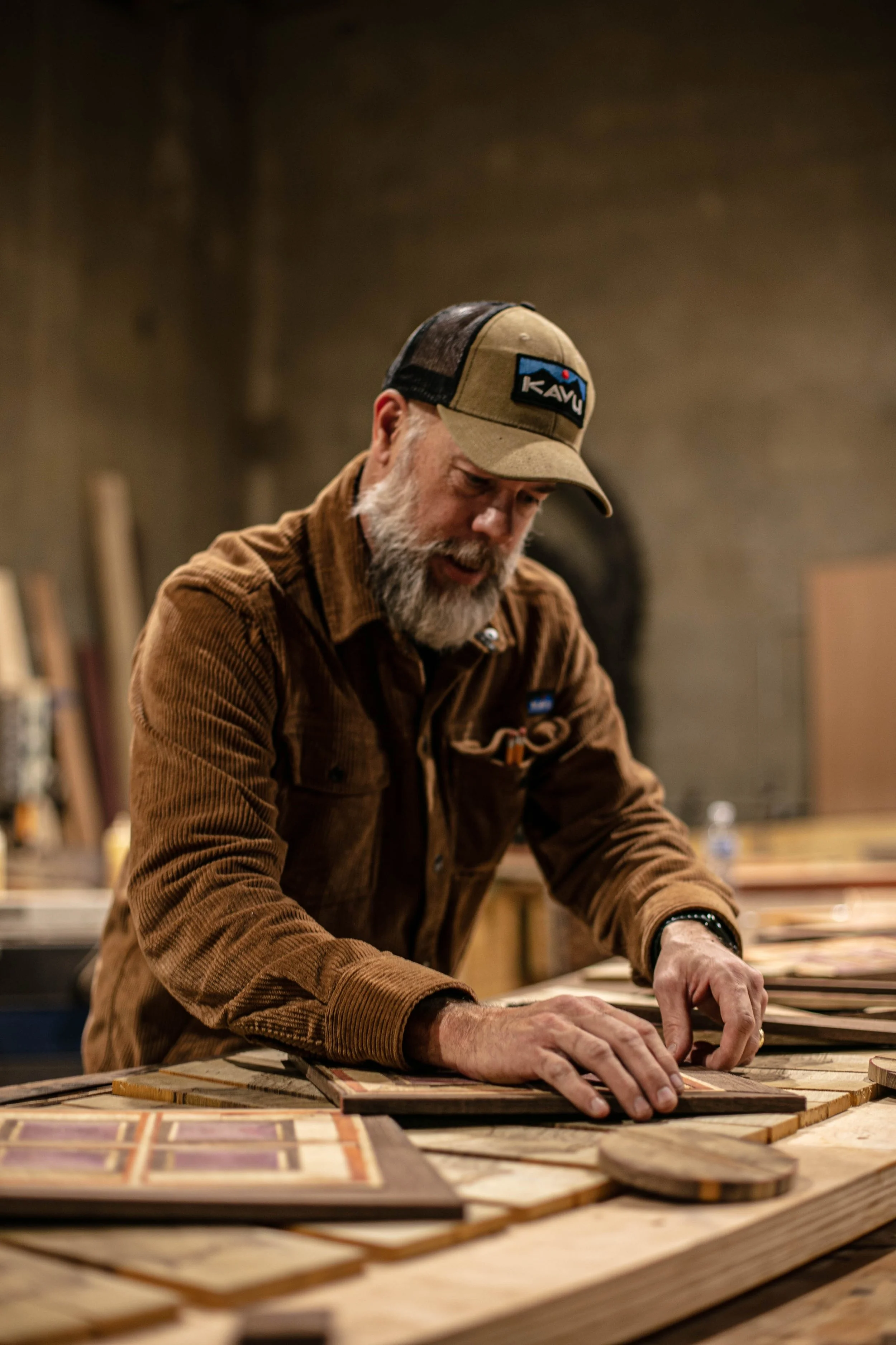 A man with a grey beard in a brown jacket and a beige cap working on a woodworking project in a workshop, surrounded by various pieces of wood and tools.