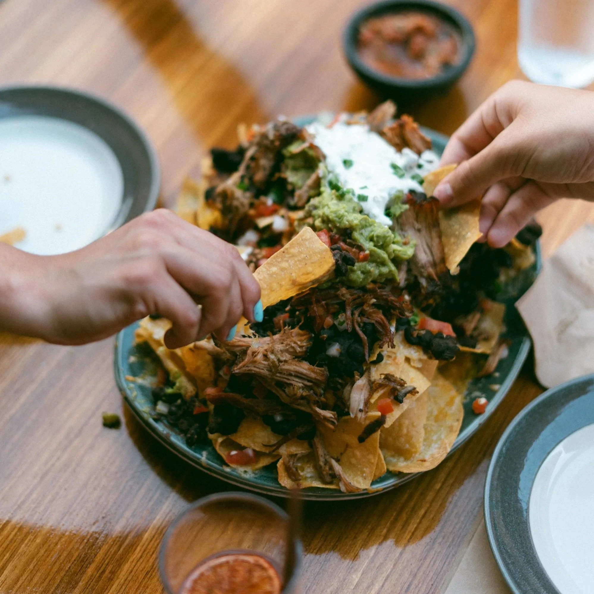 Two people sharing a large serving of nachos topped with shredded beef, guacamole, sour cream, and chopped vegetables on a blue plate at a wooden table.