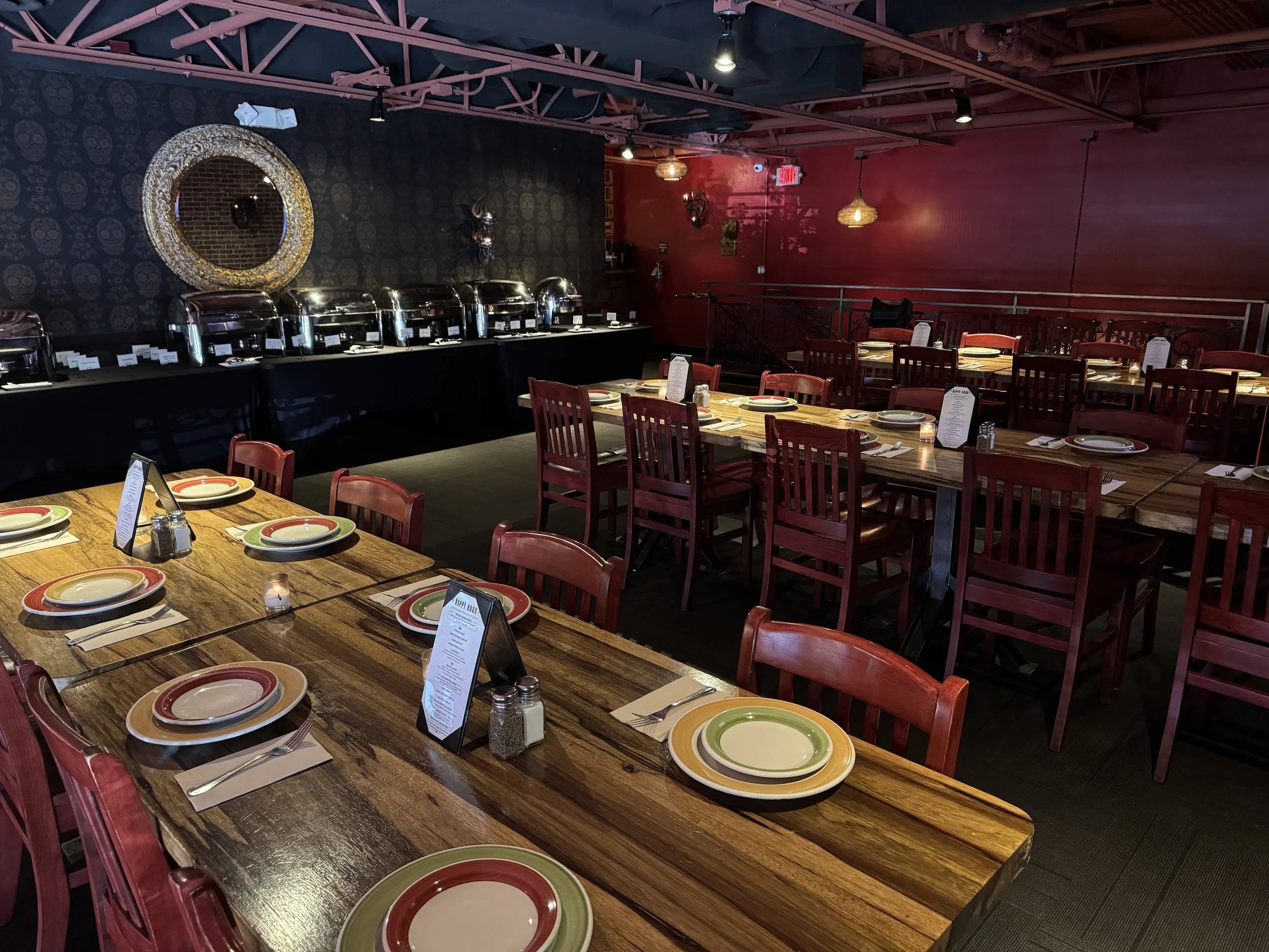 Empty restaurant with red chairs, wooden tables set with plates and silverware, and a buffet with chafing dishes against a dark wall with skull patterns.
