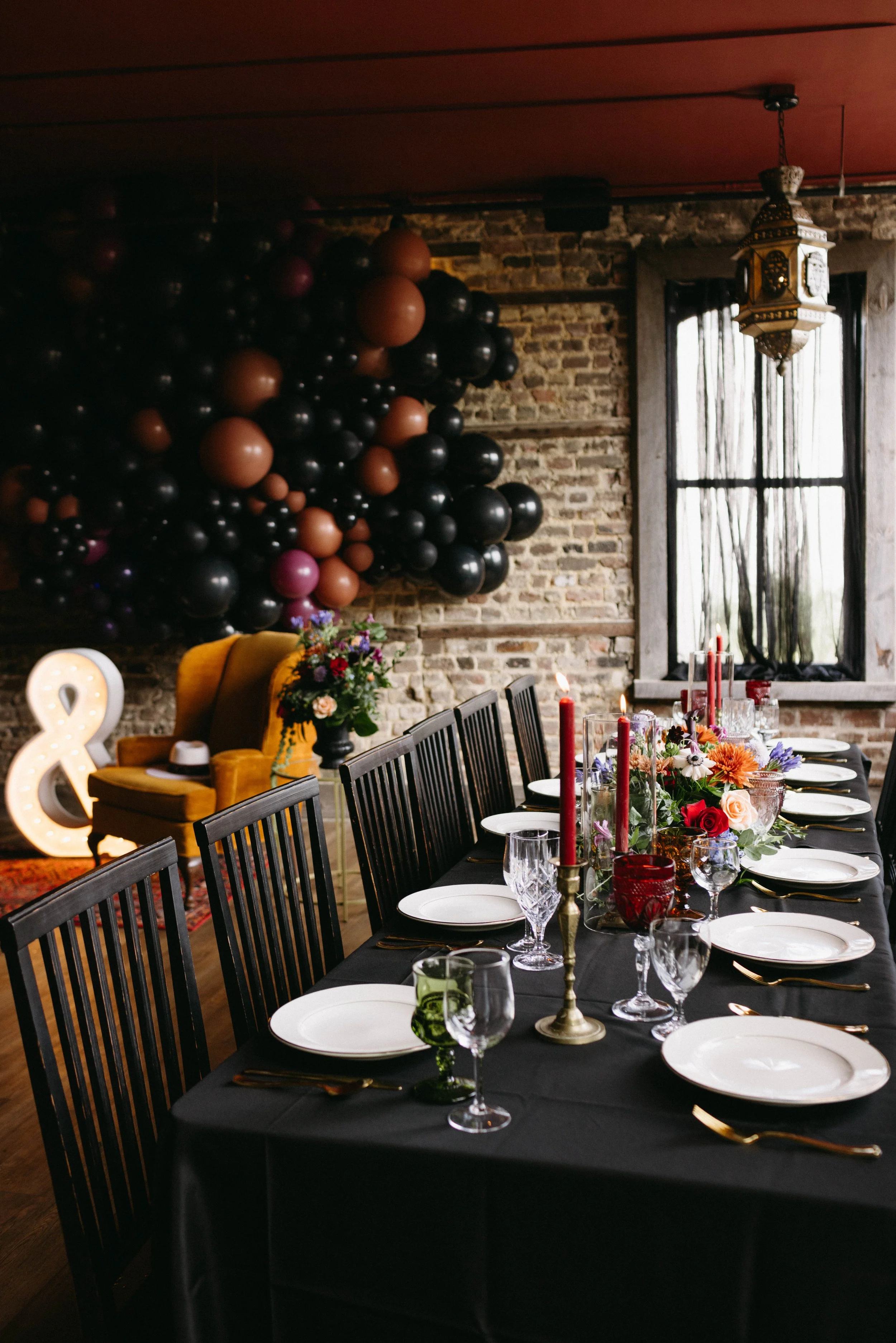 A decorated dining table set for a celebration with white plates, crystal glasses, gold utensils, red candles, and floral centerpieces, with black chairs around it, and a brick wall with black and purple balloons and a yellow armchair with a flower arrangement in the background.