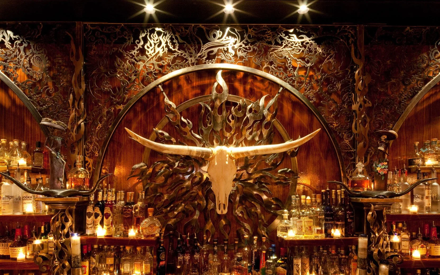 A decorative bar with a large metallic longhorn skull mounted on a wooden wall, surrounded by intricate metalwork resembling flames and animals, with bottles of alcohol and candles on shelves.