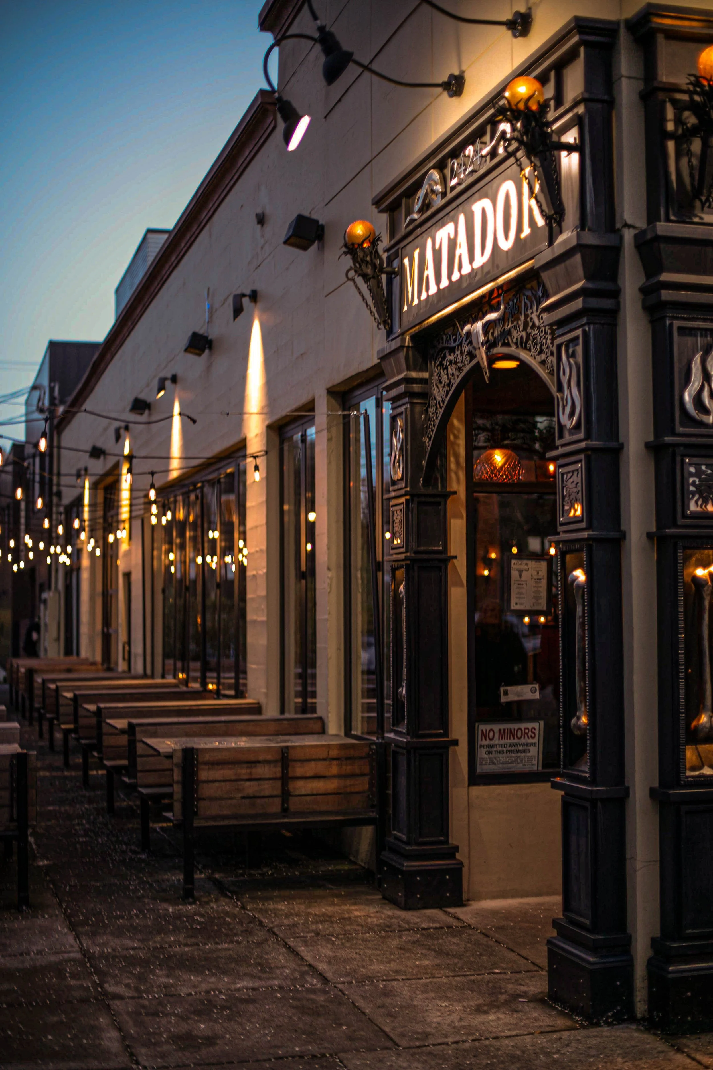 Exterior view of a restaurant named Matador with ornate black accents, warm lighting, and string lights along the sidewalk at dusk.