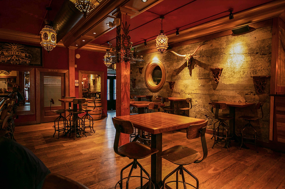 Empty rustic bar or restaurant with wooden tables and chairs, decorative lanterns, a mounted animal skull, and a textured stone wall.
