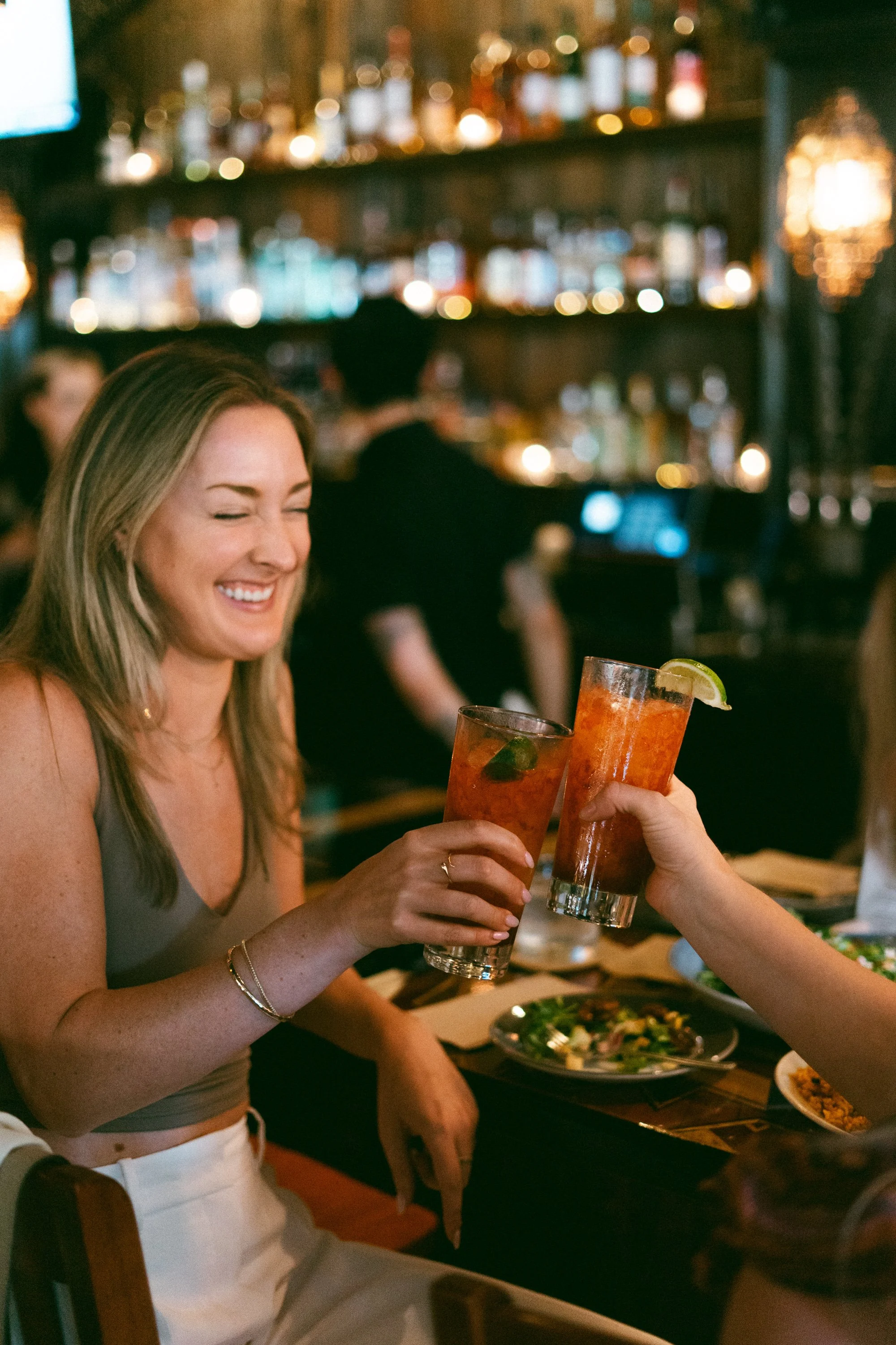 A woman smiling and laughing while toasting with drinks at a restaurant or bar.