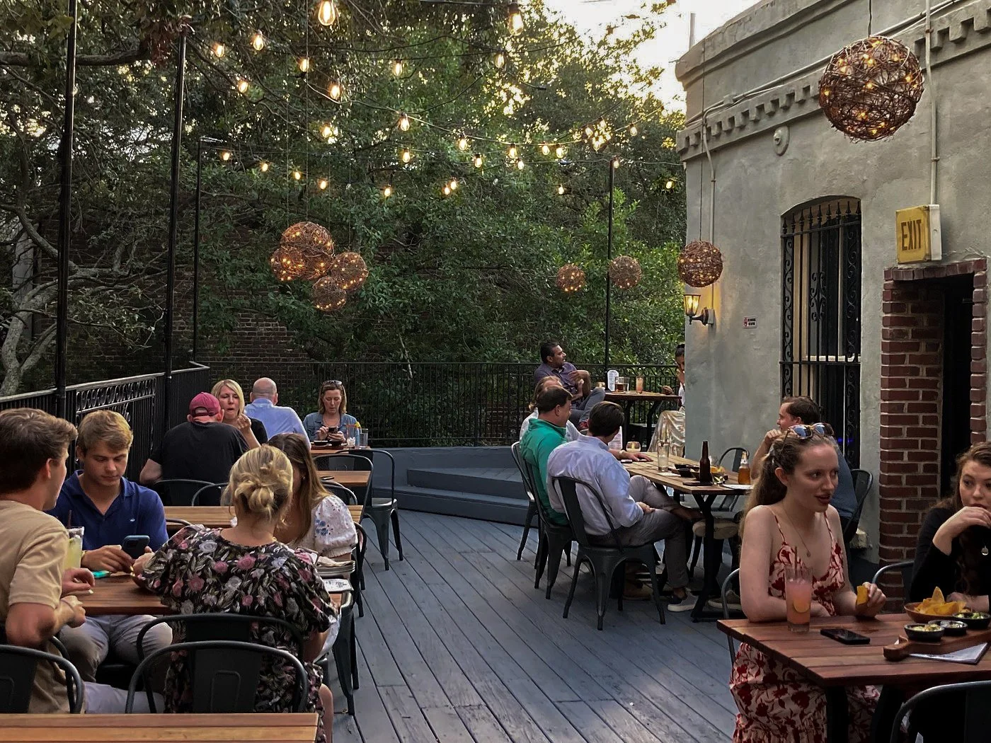 People dining on an outdoor patio with string lights and hanging spherical lanterns, surrounded by trees and a building with an exit sign.