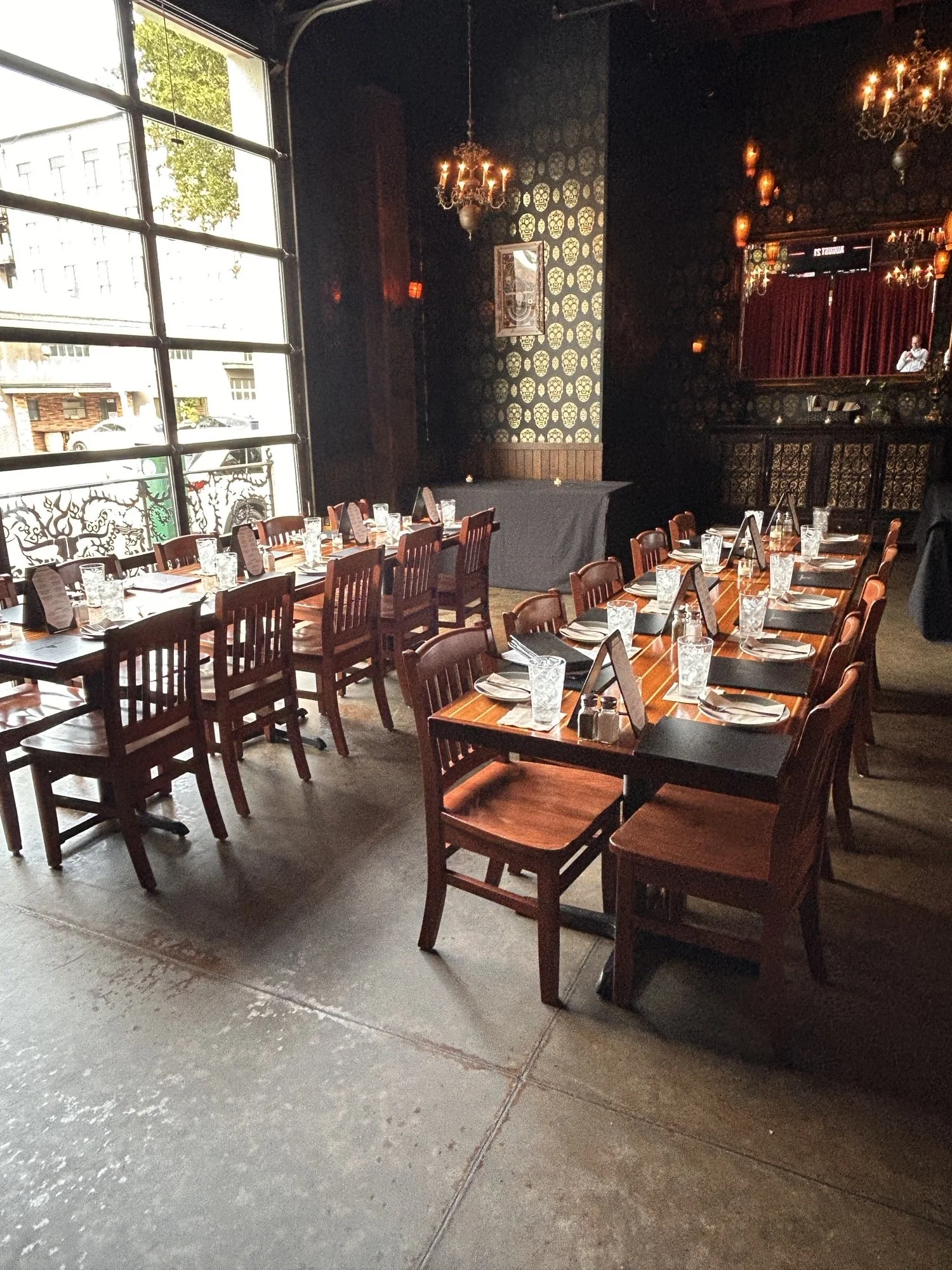 Interior of a restaurant with wooden tables and chairs, set with glasses, napkins, and plates, featuring a large window, dark wallpaper, chandeliers, and a decorated wall with mirrors.
