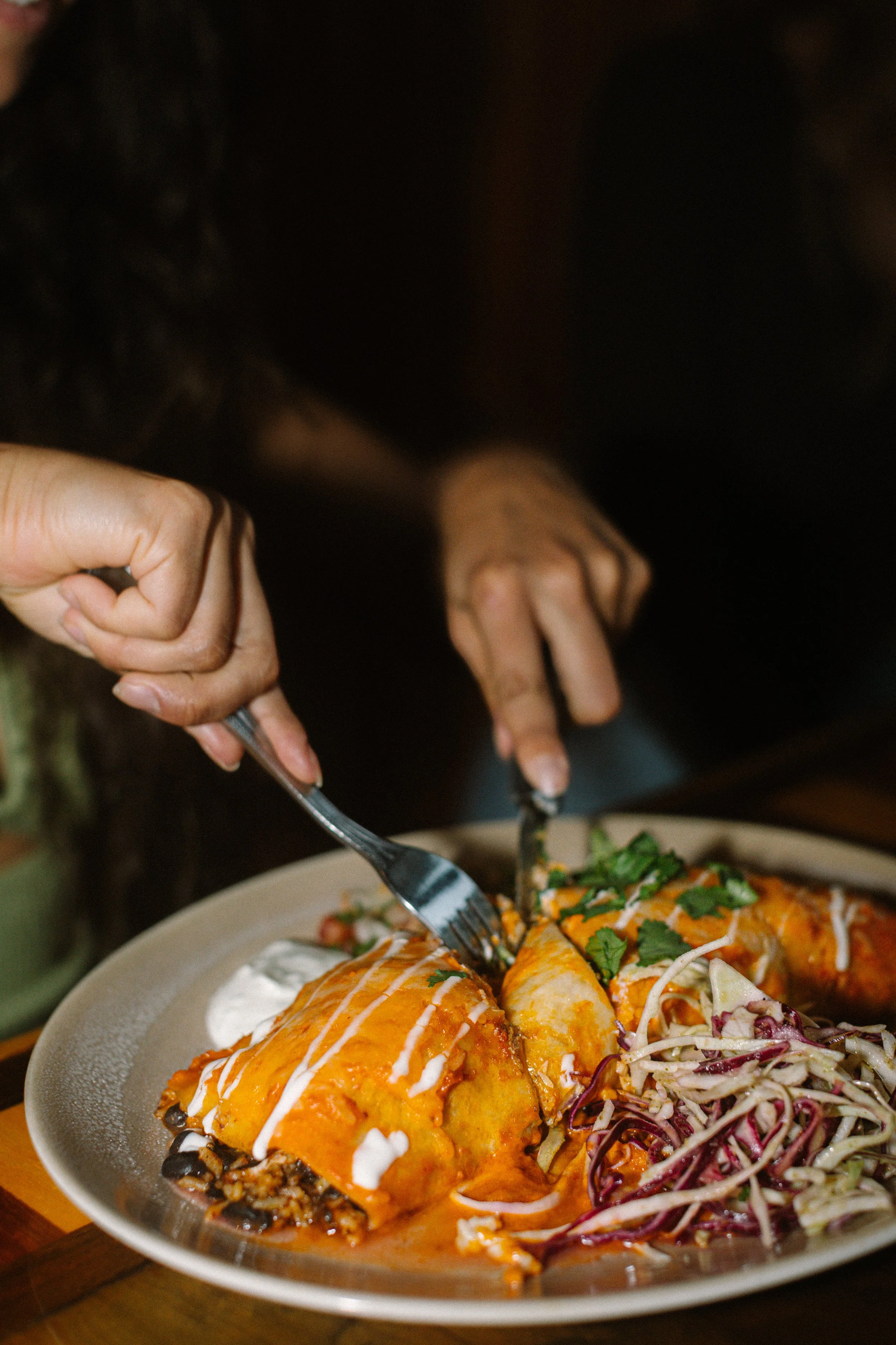 Hands using forks to cut and eat enchiladas topped with cheese and cilantro, served with a side of coleslaw on a plate.