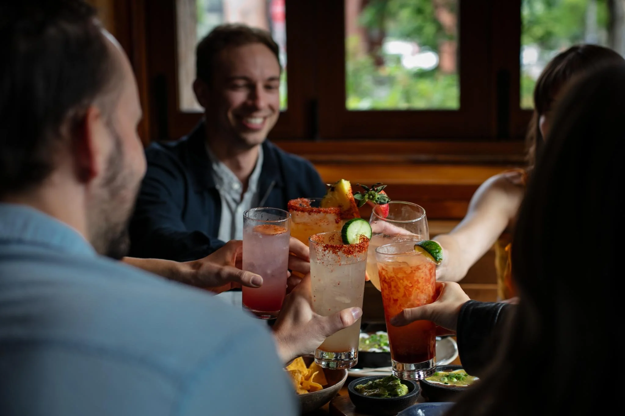 Group of four friends enjoying drinks and chips at a restaurant table, with a man smiling in the background, during daytime.