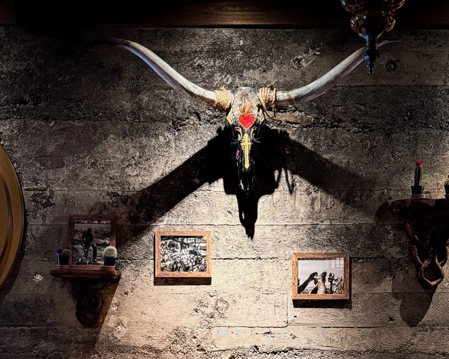 A mounted animal skull with large horns, decorated with paint and a red heart, hanging on a textured stone wall, casting a shadow. Flanking the skull are small framed black-and-white photographs, and decorative items on wooden shelves.