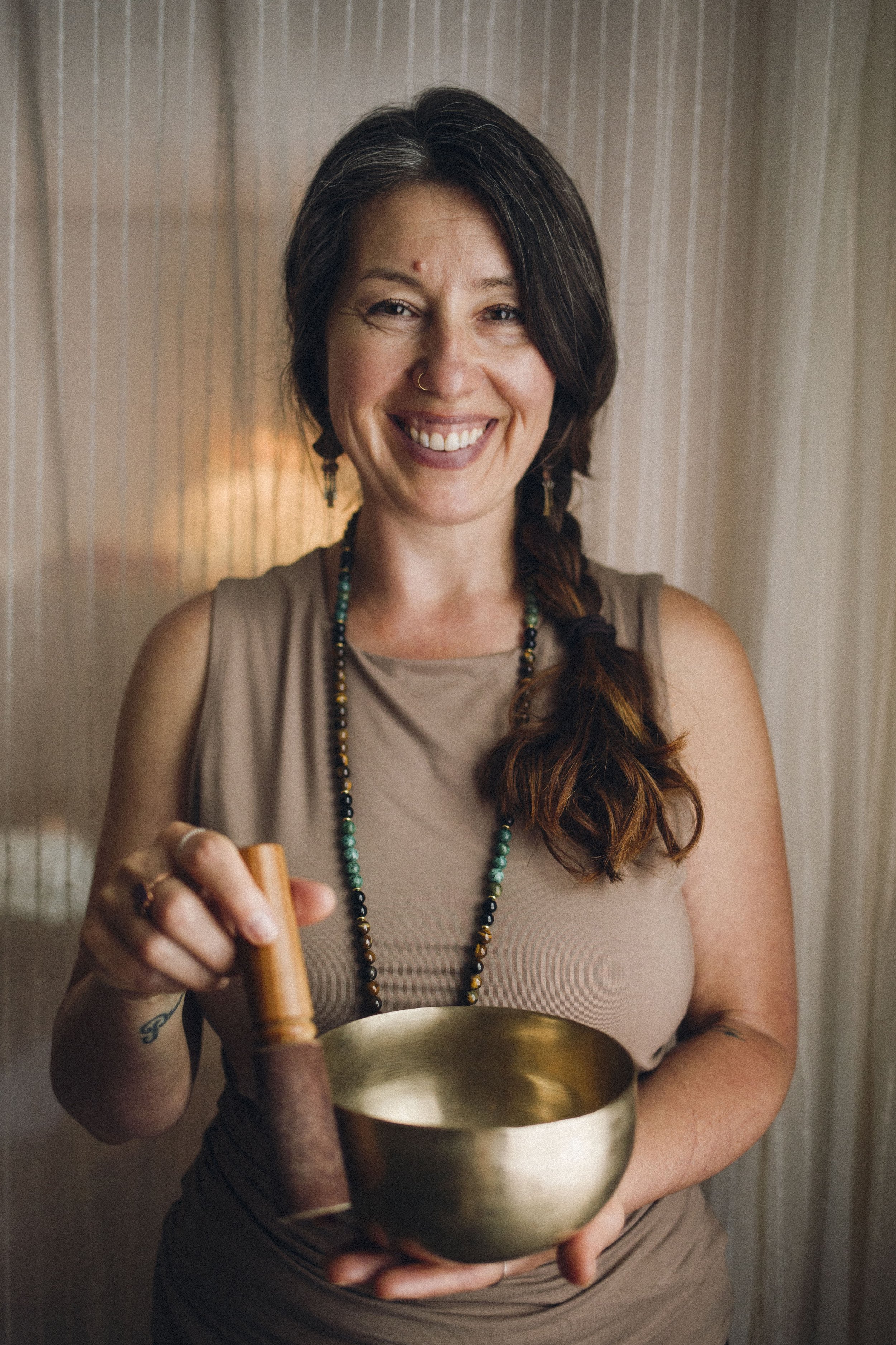 A woman with dark hair in a braid, smiling and holding a singing bowl and a mallet, standing in front of a wooden wall.  Sound healing, spiritual, nature based wisdom.  Come home to your body.