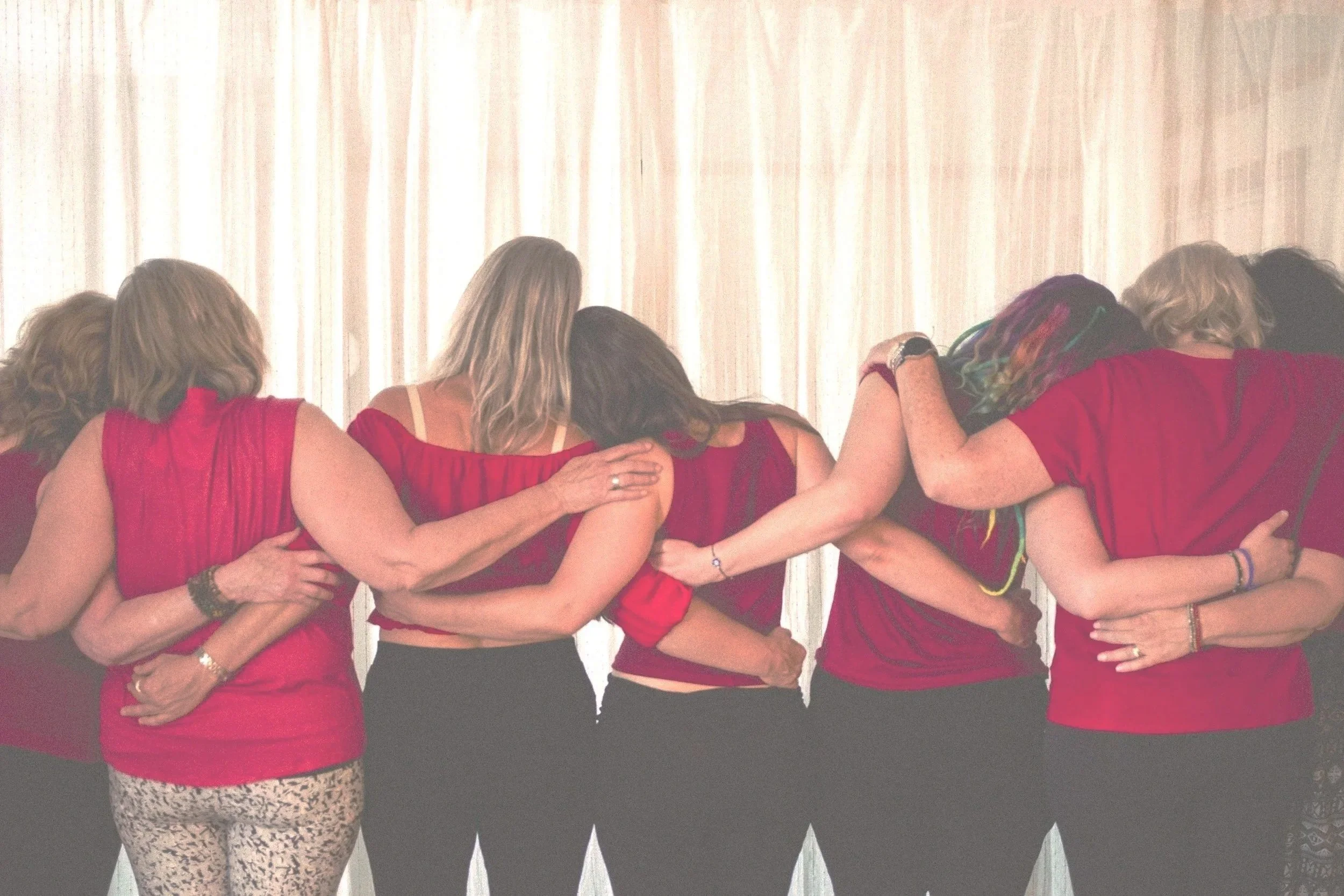 A group of women in red shirts embracing each other from behind in front of a white curtain. Community support, empowering and learning from one another.  
