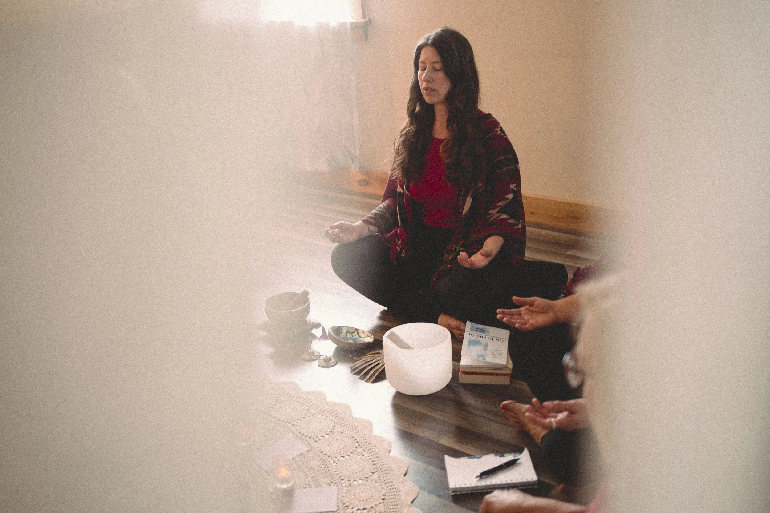 Women practicing meditation in a cozy room with spiritual items, sitting cross-legged on the floor with eyes closed and hands resting on knees. Women's circle menopause, midlife, community, spiritual, empowerment.