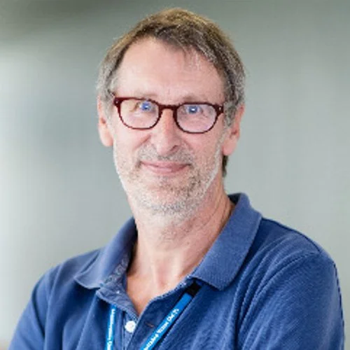 A middle-aged man with glasses, short brown hair, and a beard, wearing a blue polo shirt, standing against a light background.