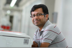 A man wearing glasses and a striped polo shirt sitting at a desk or table in a bright indoor setting.