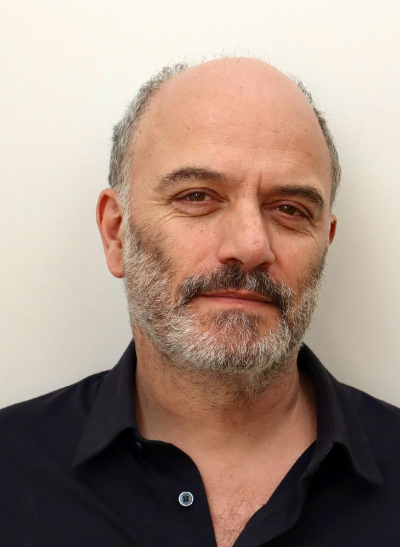Close-up of a middle-aged man with a beard, smiling slightly, wearing a black shirt, standing against a plain light-colored background.