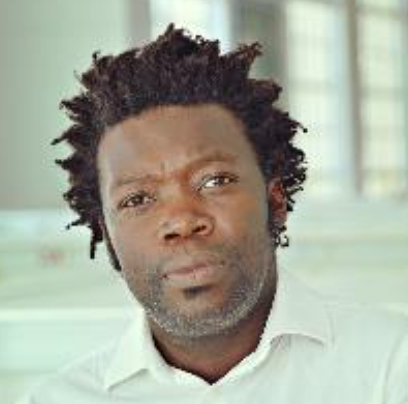 Close-up of a man with dark, curly hair, a beard, and a serious expression, wearing a white shirt.