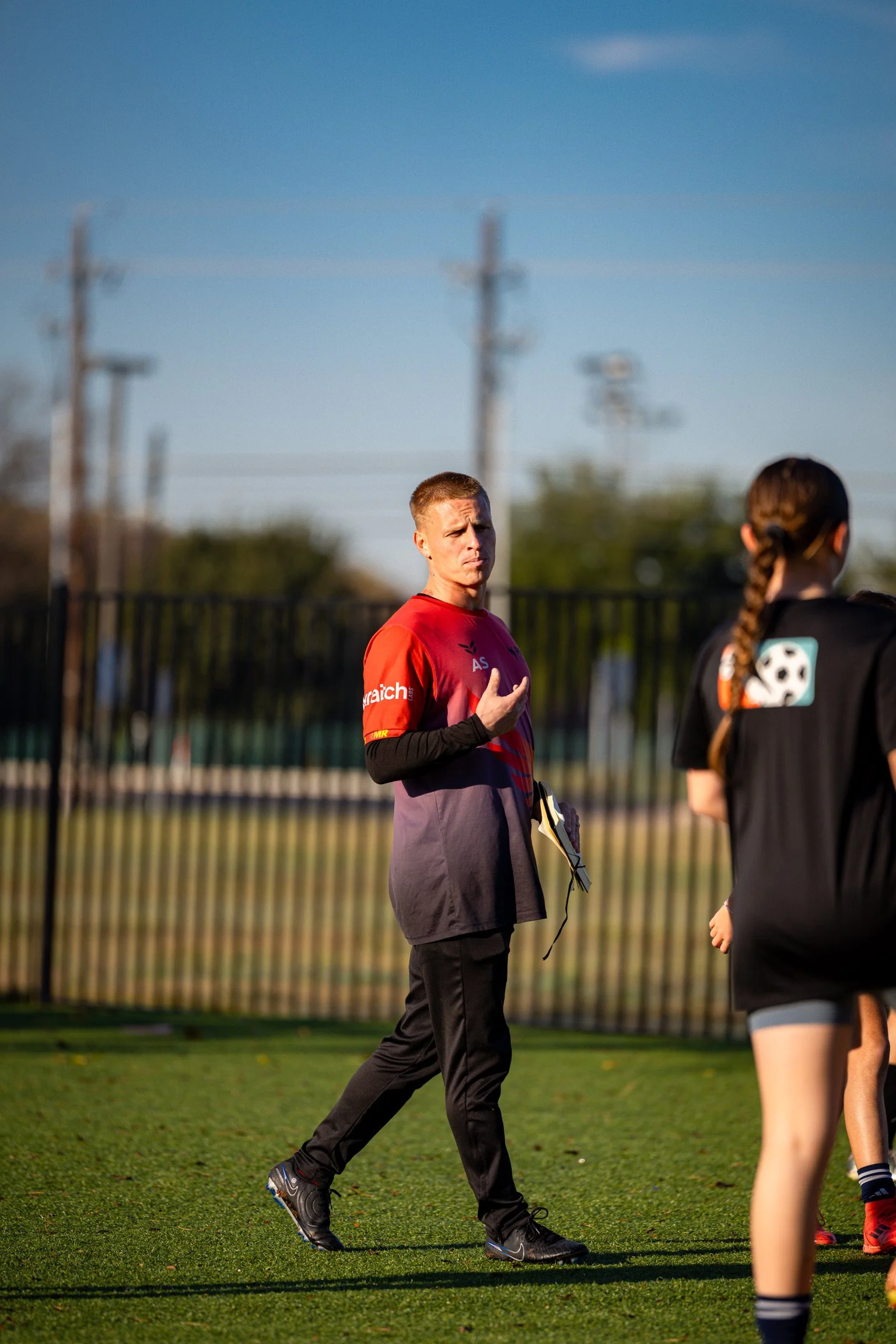 A male coach in a red shirt and black pants speaks to a group of female soccer players on a field during late afternoon or early evening.