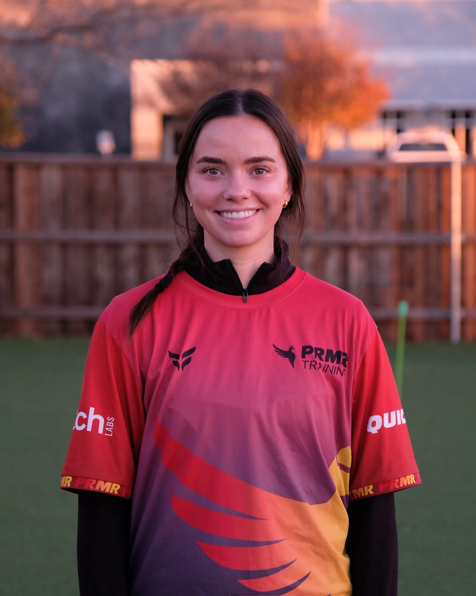 A young woman with dark hair in a braid, smiling while standing outdoors during sunset. She wears a pink sports jersey with logos and a black zip-up underneath. Background features a wooden fence, trees with orange leaves, and parked cars.
