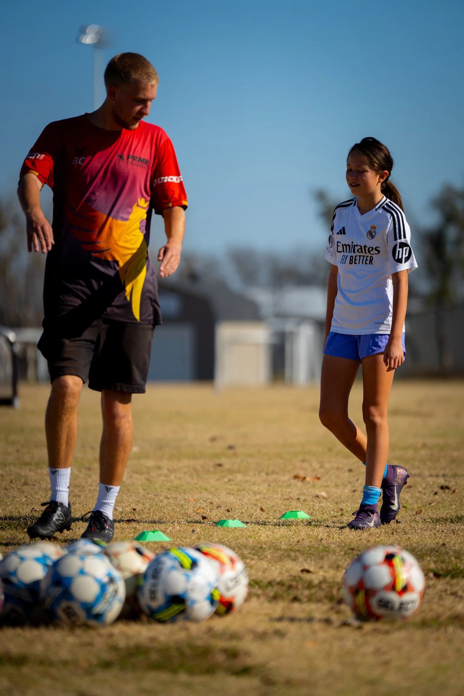 A young girl and a man, likely a coach, standing on a grassy field with soccer balls and small orange cones, preparing for a soccer training session on a clear day.