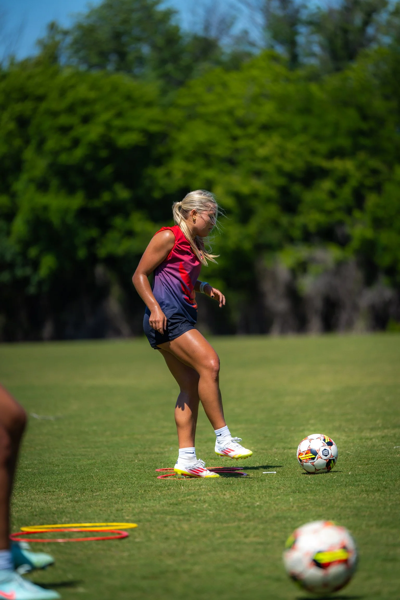 A female soccer player in a red and purple jersey practicing dribbling with a soccer ball on a grass field with trees in the background.